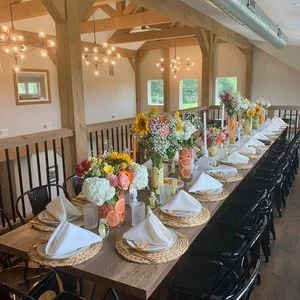 Long wooden dining table set for a gathering with white plates, napkins, and woven place mats. The table features colorful floral centerpieces with sunflowers, roses, and other flowers. Overhead, string lights hang from the wooden beams, creating a w