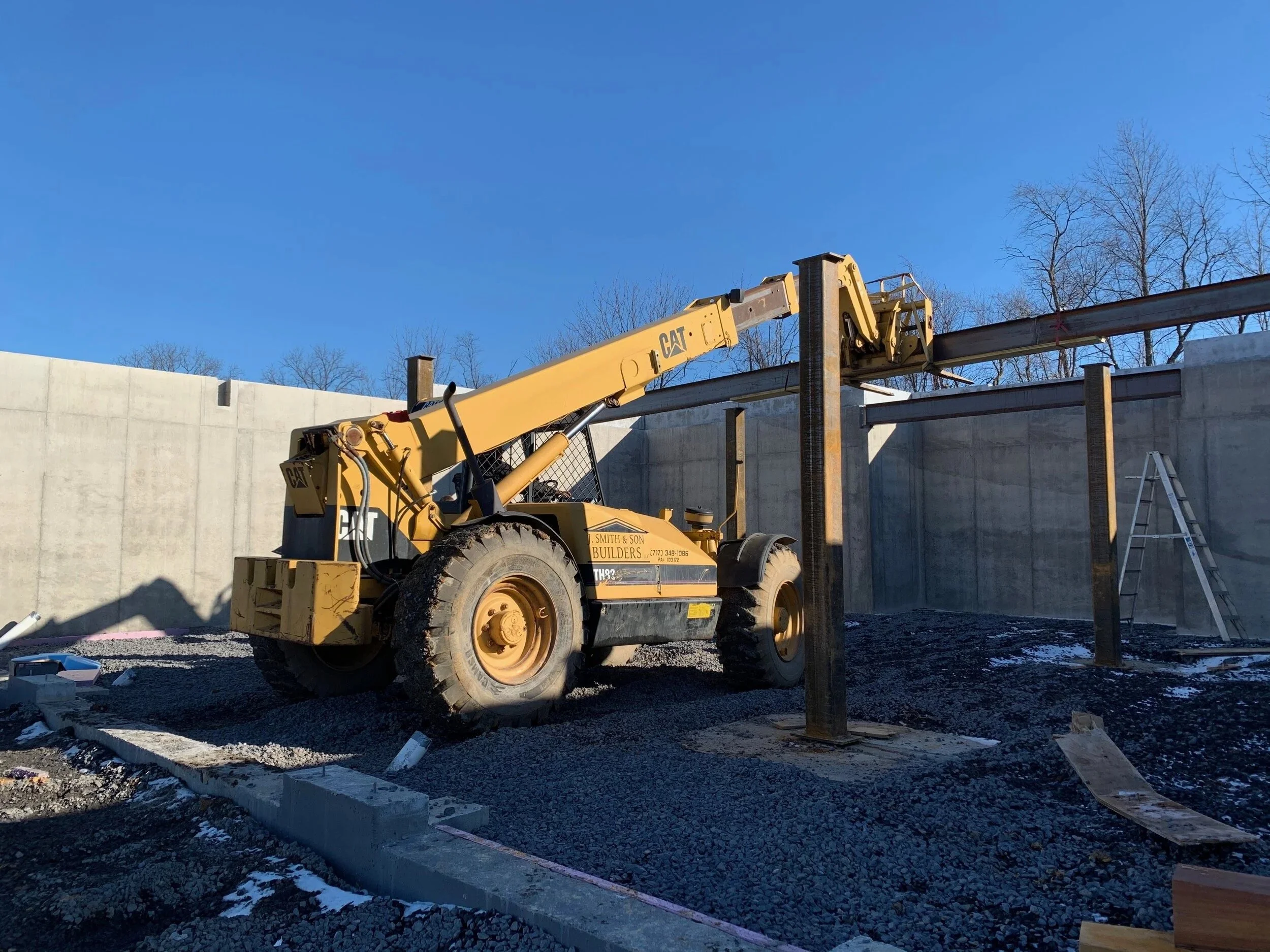 Construction site with a yellow CAT telescopic boom lift working on a concrete wall, ladder leaning against the wall, clear blue sky, and trees in the background.