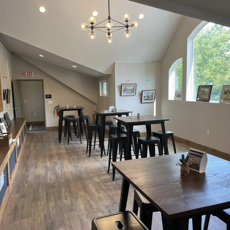 Interior of a bright, modern cafe with high vaulted ceiling, large window showing green trees outside, wooden floor, black tables and chairs, framed artwork on walls, and a decorative chandelier hanging from the ceiling.
