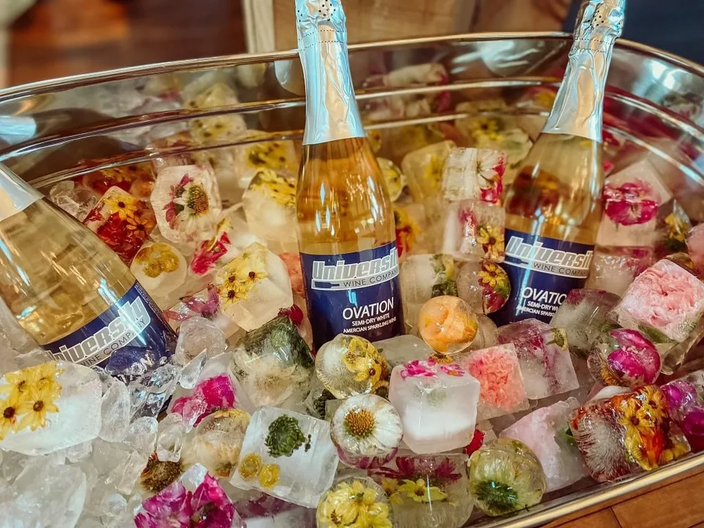 Three bottles of Unwersal Ovation semi-dry white wine in a bowl filled with ice cubes and decorative edible flowers.