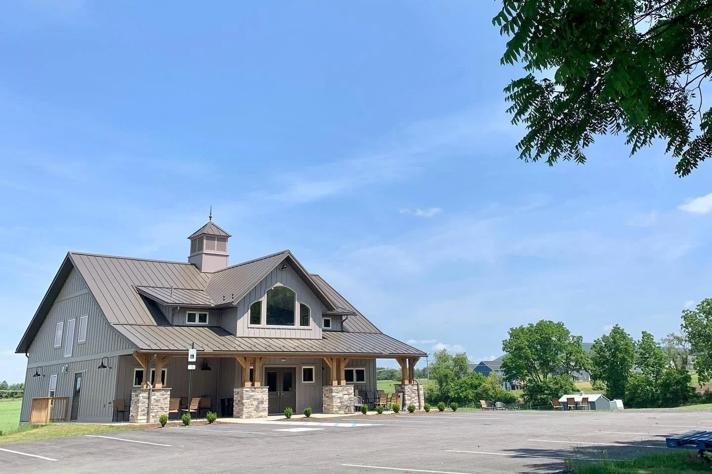A gray, barn-style building with a metal roof, stone accents, a small porch, parking lot, and trees in the background under a clear blue sky.