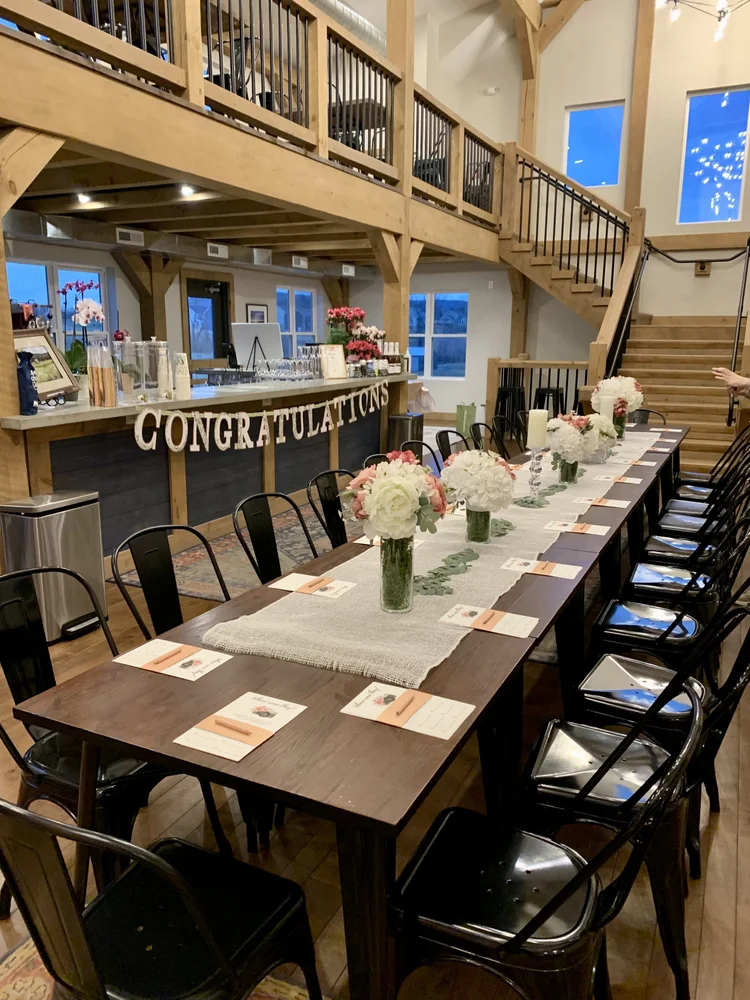 A decorated indoor dining area for a celebration, with a long table set with floral centerpieces, place cards, and a banner saying 'CONGRATULATIONS' in the background.