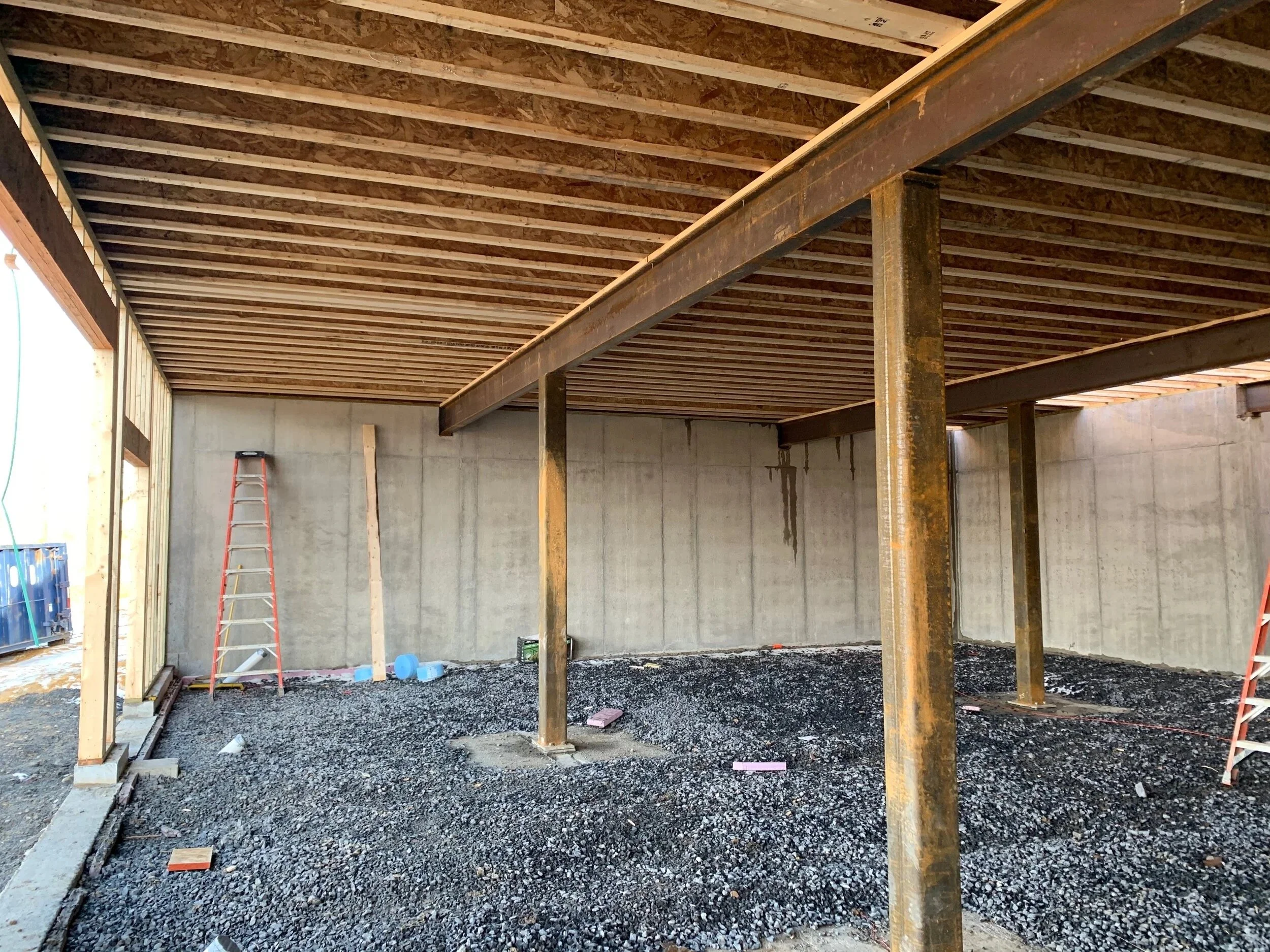 Interior view of a building under construction showing a concrete foundation, steel beams, and exposed wooden framing on the ceiling. Construction tools and materials are visible on the gravel floor.