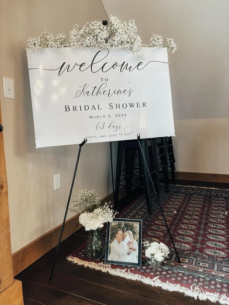 Welcome sign for Katherine's bridal shower on March 2, 2024, with flowers, a framed photo of a couple, and stacked black chairs in the background.