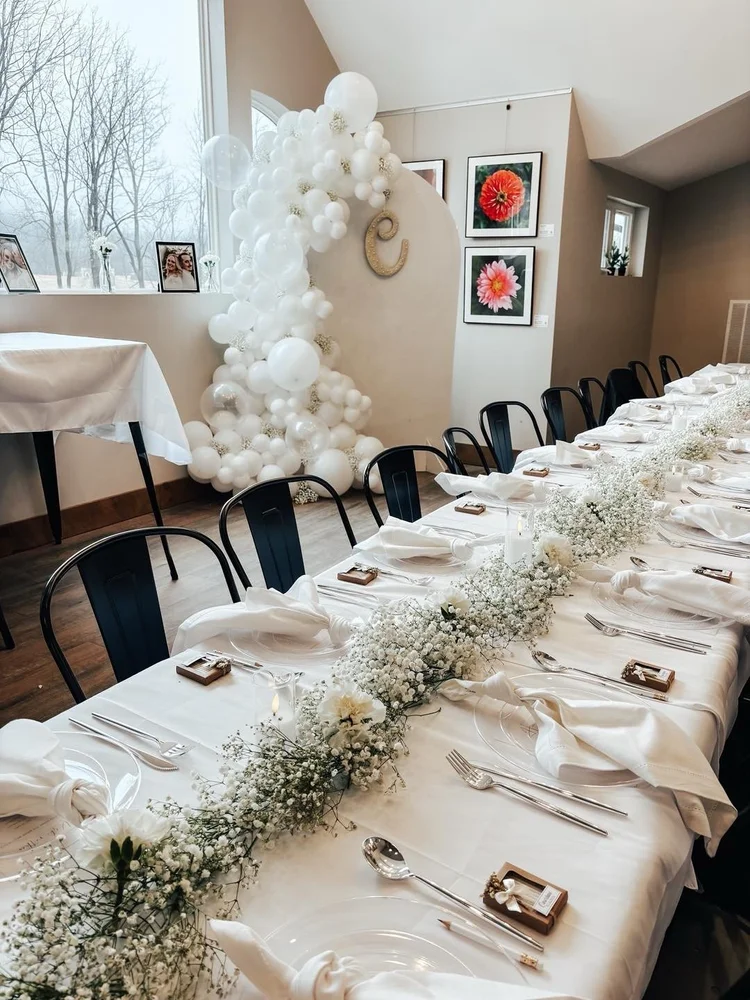 Long banquet table decorated with white tablecloth, white napkins, floral centerpieces, and place settings in a bright room with balloon and flower wall decor.