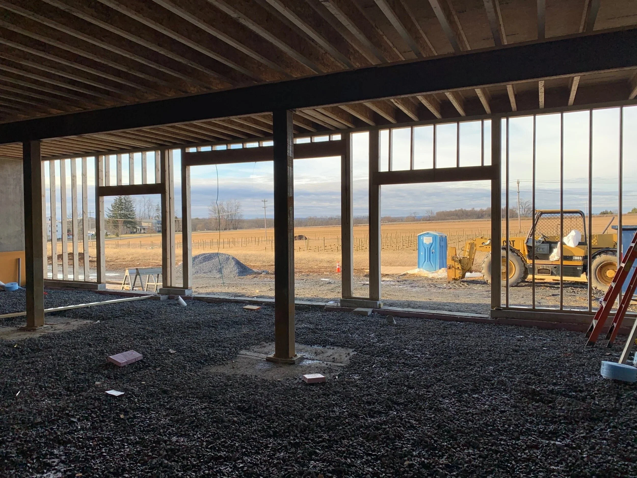 Construction site of a building with framing in progress, view from inside looking out at a rural landscape with a yellow bulldozer, blue portable toilet, and open field.