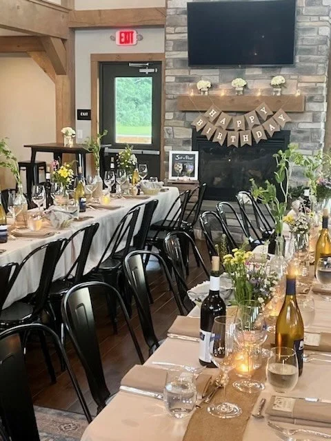 Decorated indoor party table with floral centerpieces, wine bottles, wine glasses, candles, and place settings, with a 