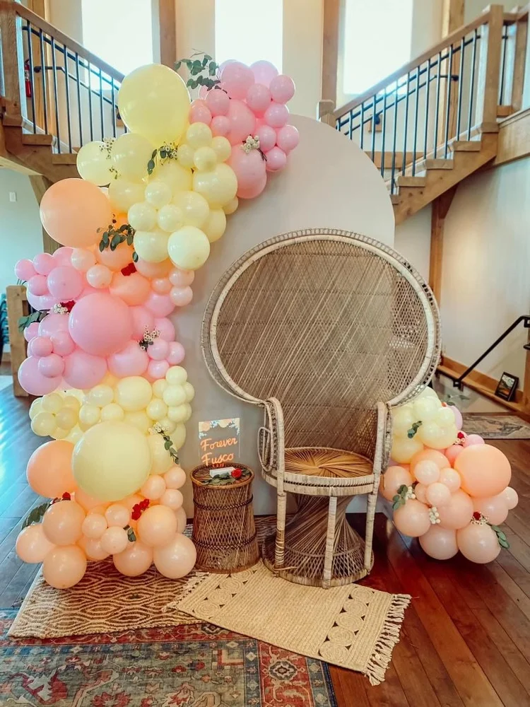 Decorative arrangement with pink, yellow, and peach balloons, some greenery, a rattan chair, a small side table with a sign that says "Forever Fusco," and a woven rug, set in a spacious room with a staircase and wooden floors.