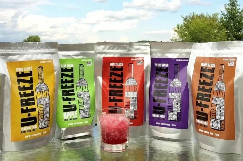 Five juice pouches with different flavors, arranged in a row outdoors on a reflective surface with a blue sky and green trees in the background, and a glass of berry juice in front.
