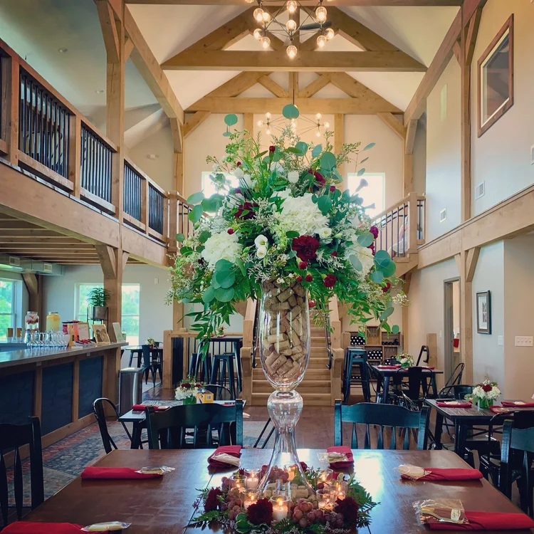 A large flower arrangement with white, red, and purple flowers and green foliage in a tall glass vase on a dining table in a rustic event space with wood beams and high ceilings.