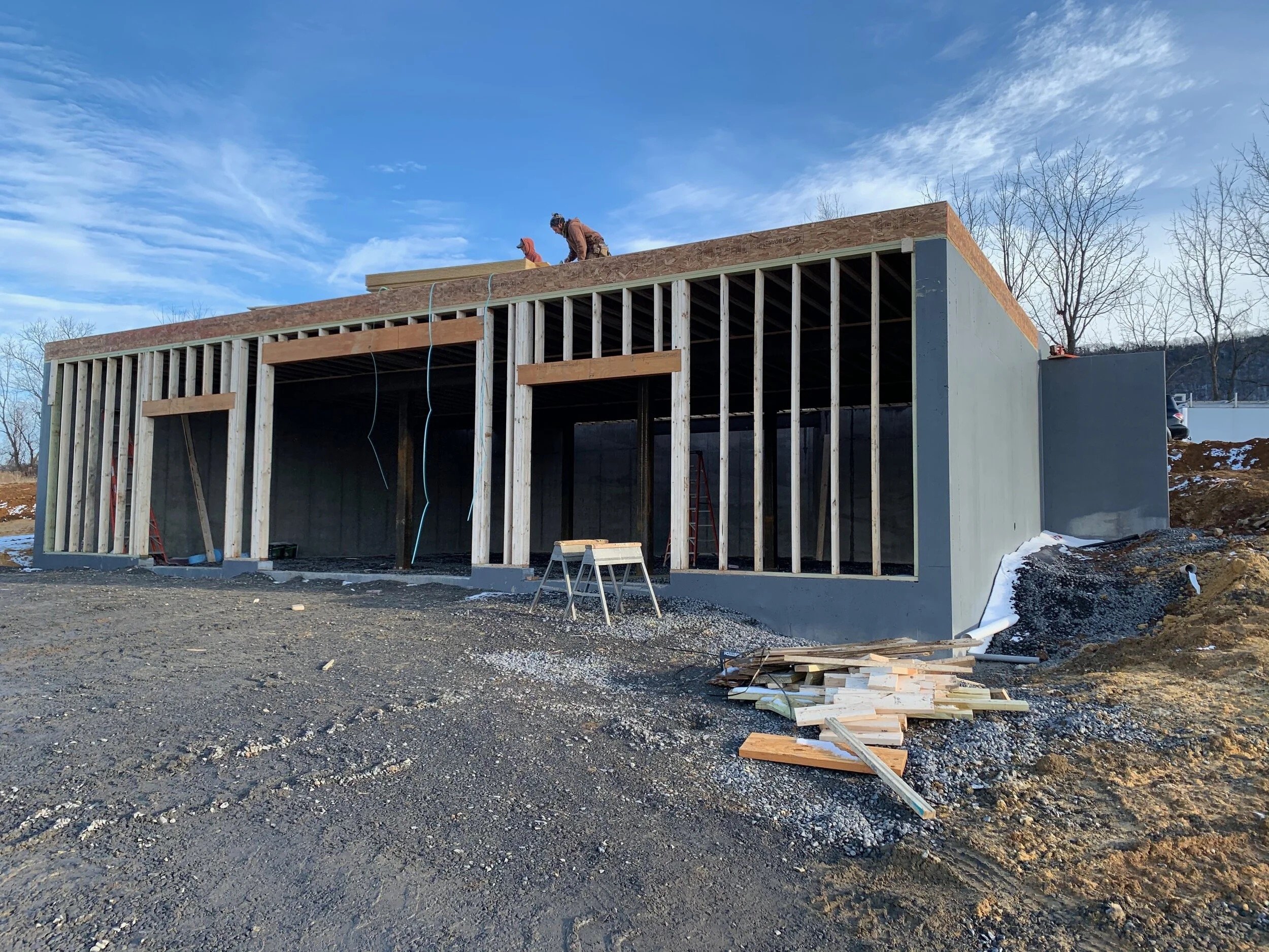 Construction site showing a building under construction with wooden framing, concrete foundation, and two workers on top of the structure. Piles of wood and construction materials are on the ground.