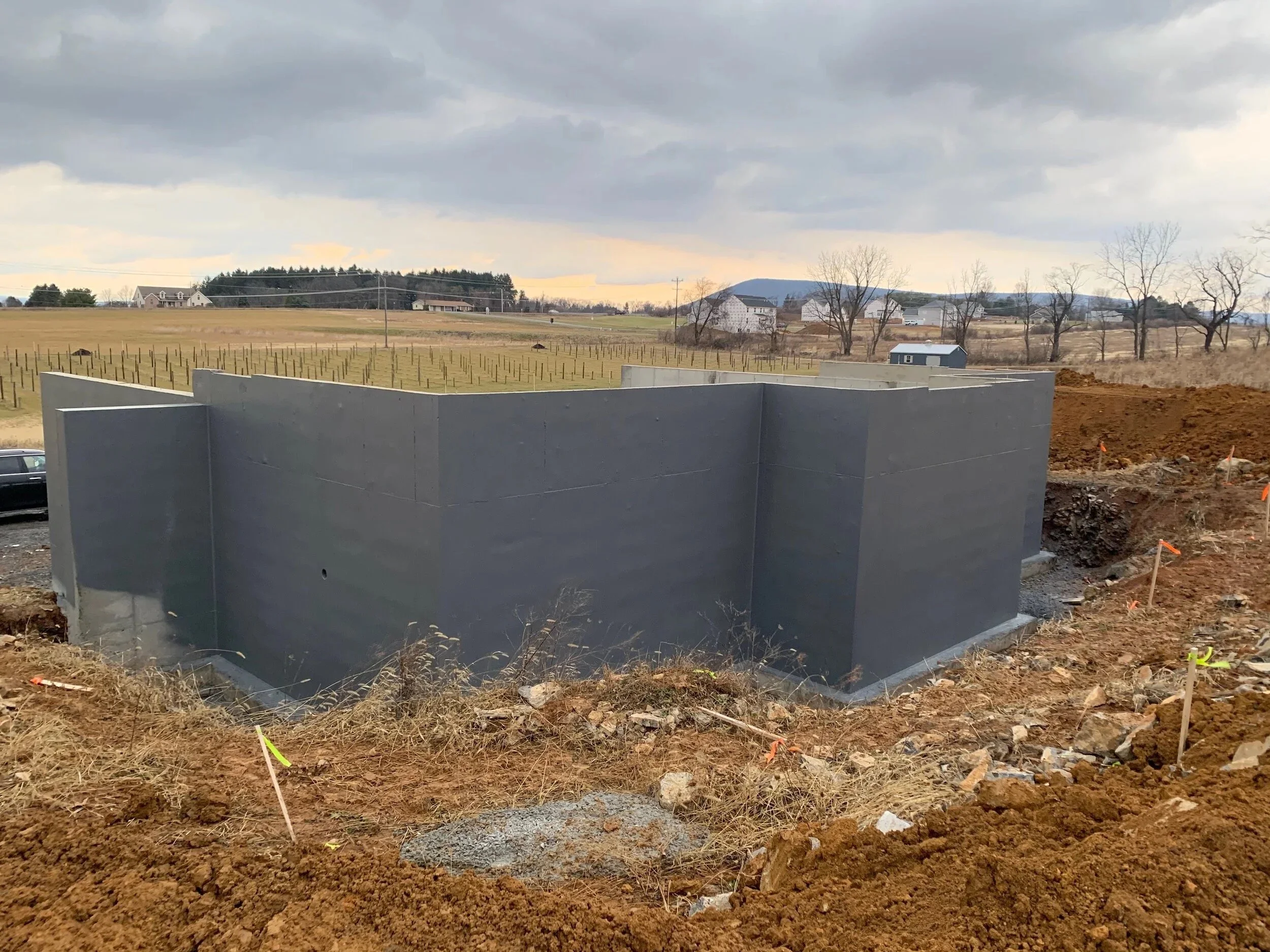 Construction site with a large, gray concrete foundation wall in a rural area, with a grassy field, trees, and houses in the background under a cloudy sky.