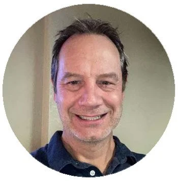 Smiling man with dark hair and a five o'clock shadow in front of a neutral wall.