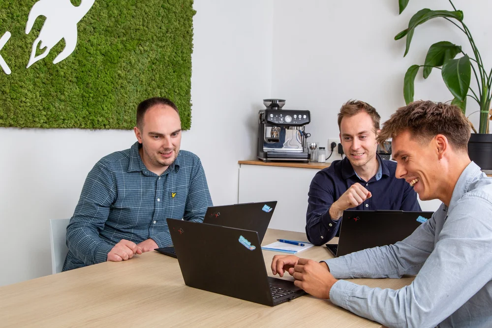 Three colleagues collaborating around laptops at a desk, greenery in background