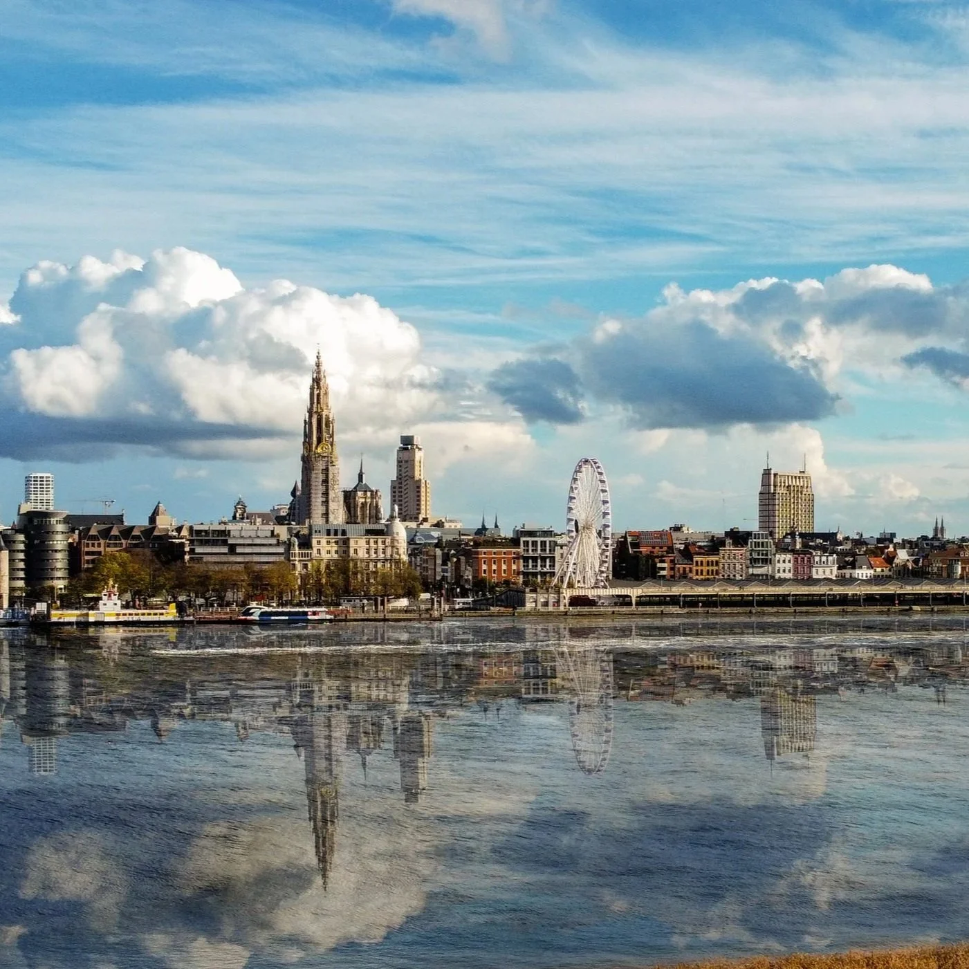 Antwerp city skyline with historic buildings, a Ferris wheel, and skyscrapers reflected in a river against a partly cloudy sky.