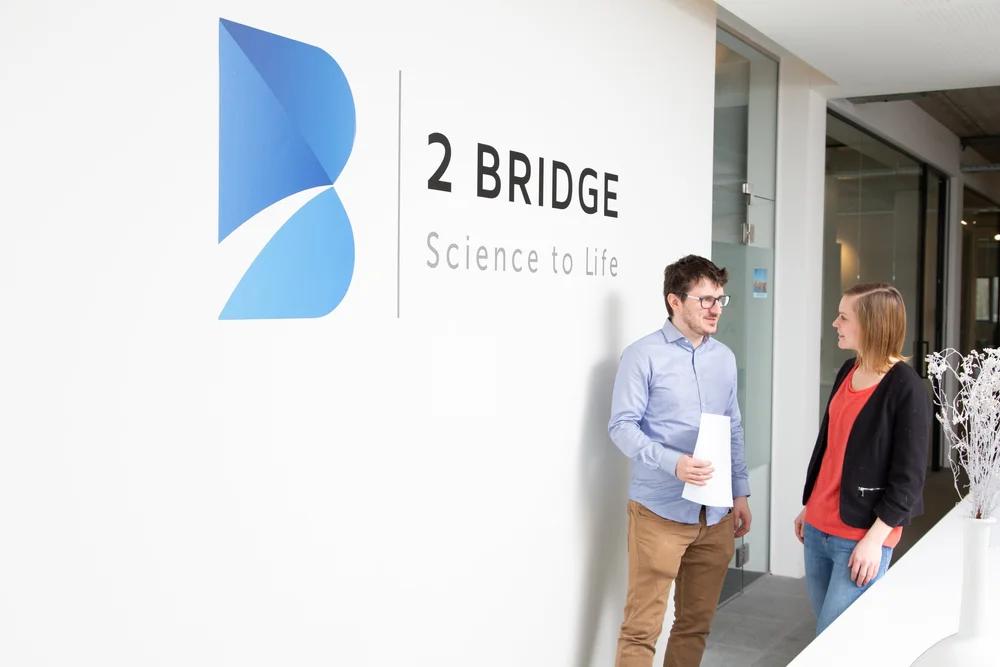 Man walking past a wall sign reading '2 BRIDGE — Science to Life'