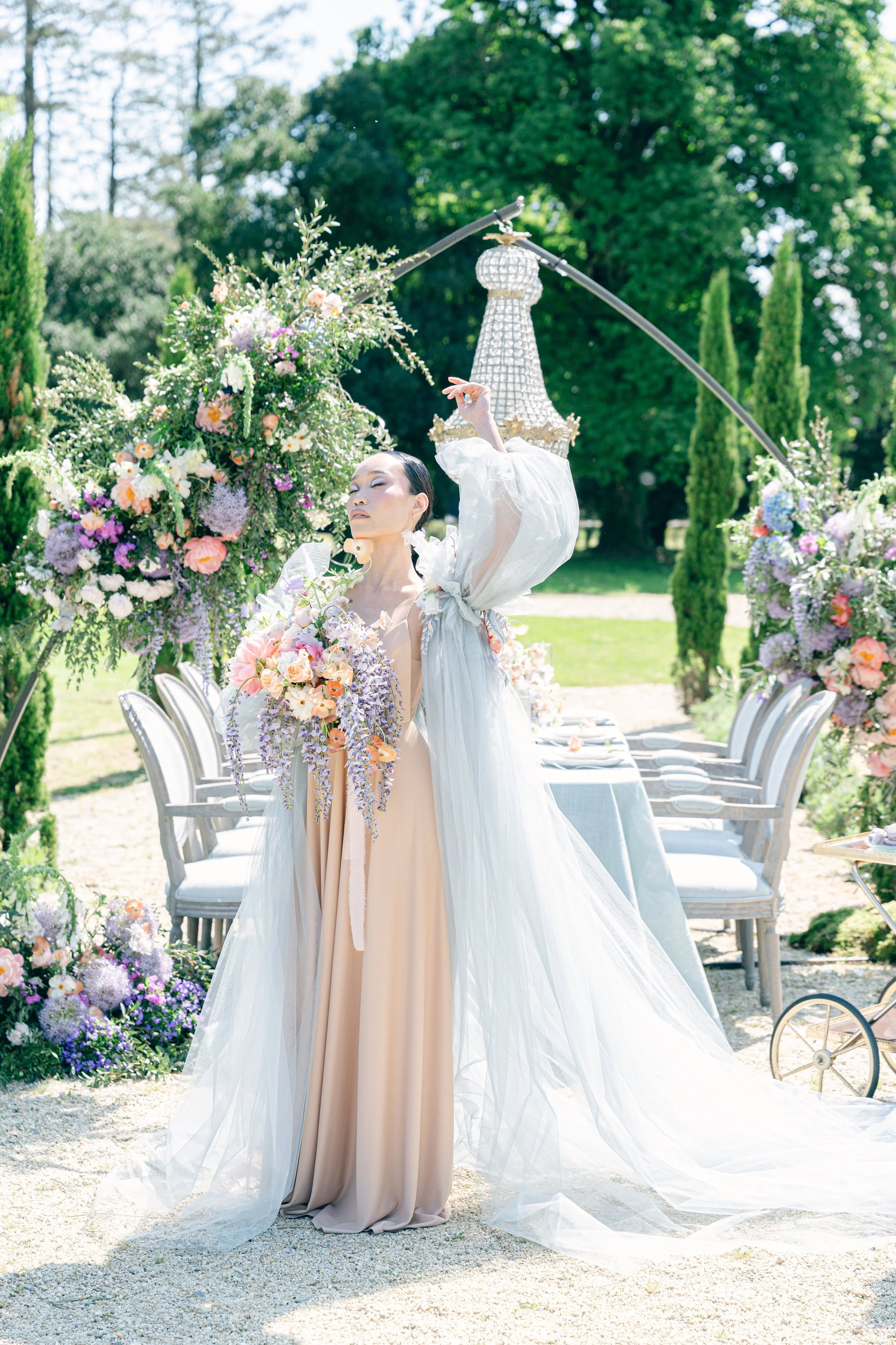 Mariée moderne tenant un bouquet de mariée cascade dans le parc d’un château en Anjou, lors d’un dîner de mariage en extérieur, élégant et fleuri, en été.