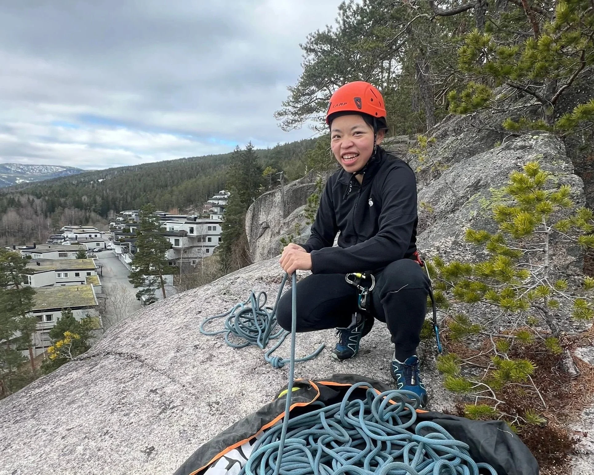 Klatreinstruktør smiler til kameraet, mens hun sitter på toppen av en klippe i drammen og forbereder en rappell.