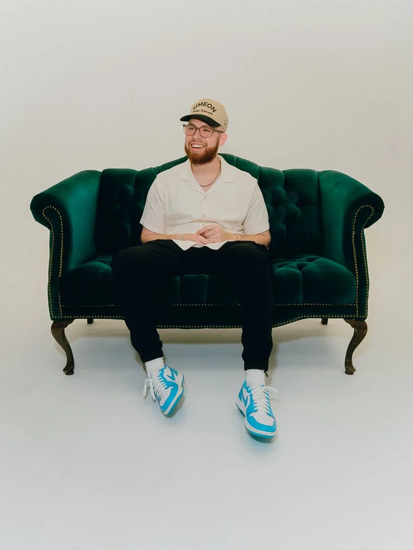 A young man with glasses and a beard, sitting on a green velvet sofa, smiling, wearing a beige cap, white shirt, black pants, white socks, and blue and white sneakers, against a plain light-colored background.