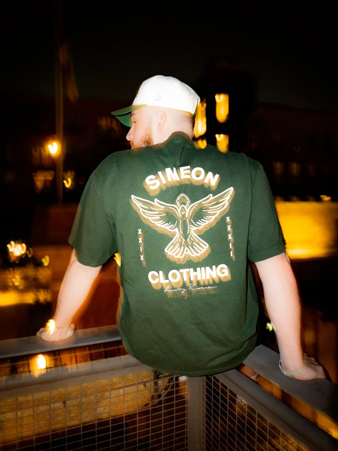 A man sitting on a railing at night, wearing a black T-shirt with "SIMEON CLOTHING" and a graphic of a flying bird on the back, and a white cap with a green brim.
