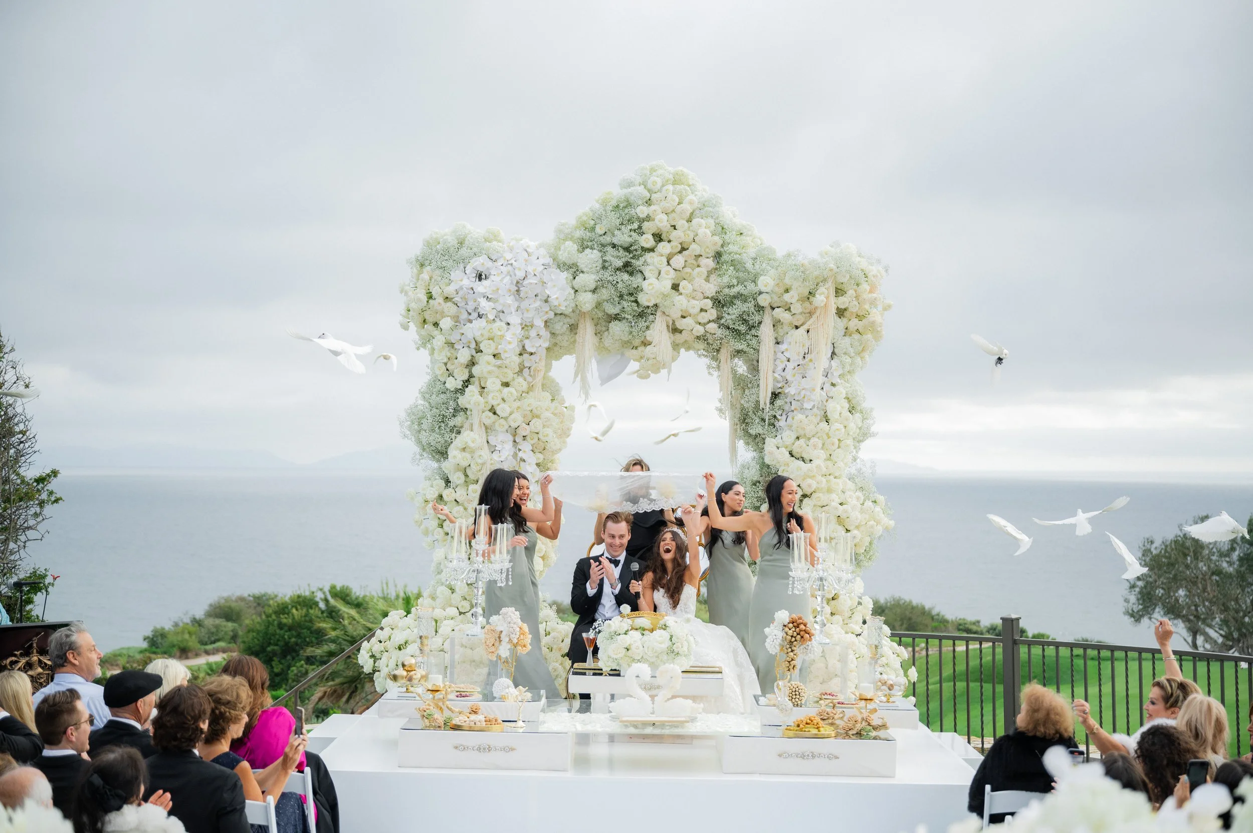 Outdoor wedding ceremony by the ocean, featuring a couple under a decorative floral arch with white roses and greenery. Attendants hold a lace canopy over the couple. Doves are in flight around the scene, while guests are seated watching the event.