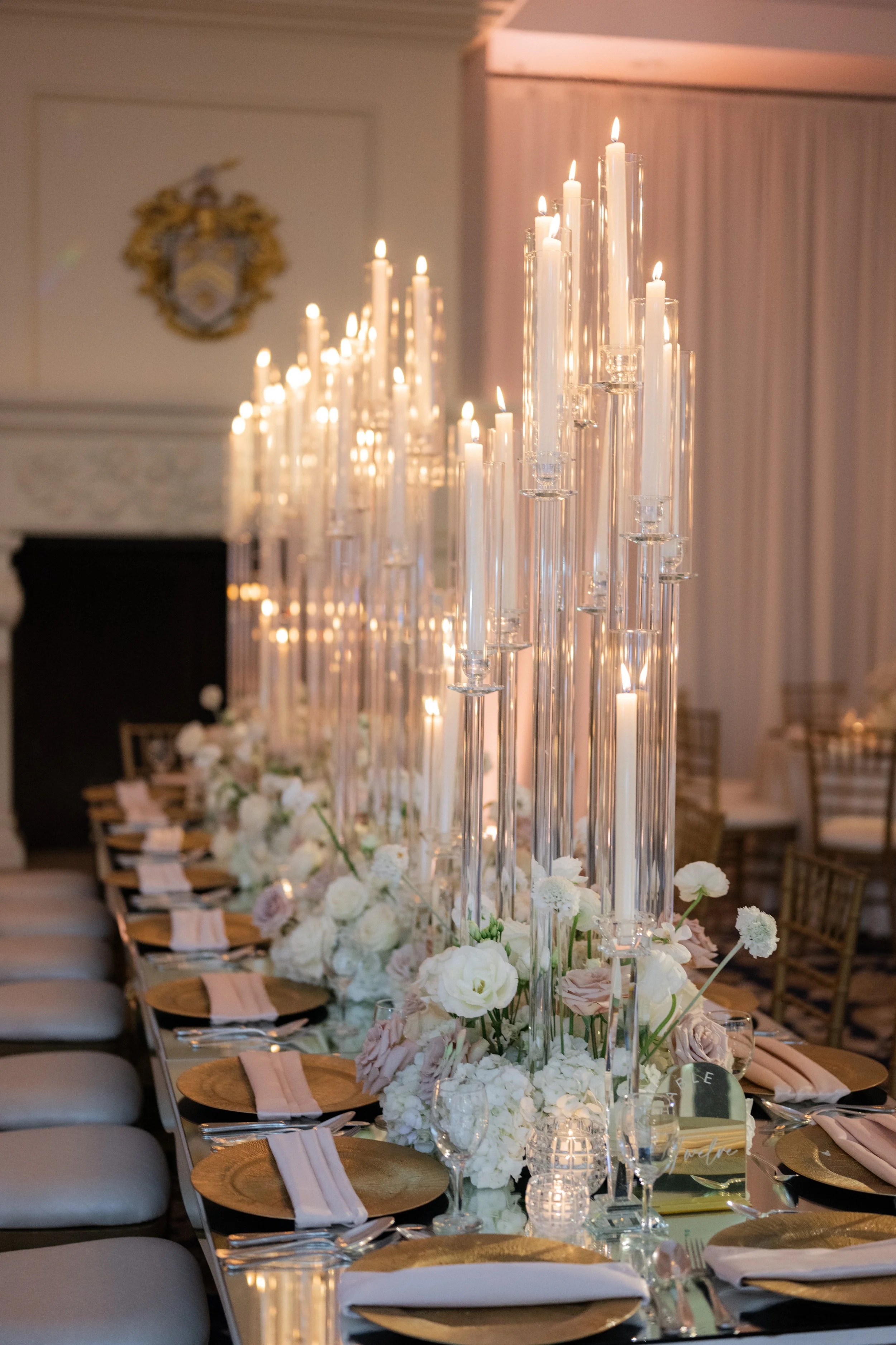 Elegant dining table setup with tall glass candle holders, white flowers, gold chargers, and neatly arranged napkins.