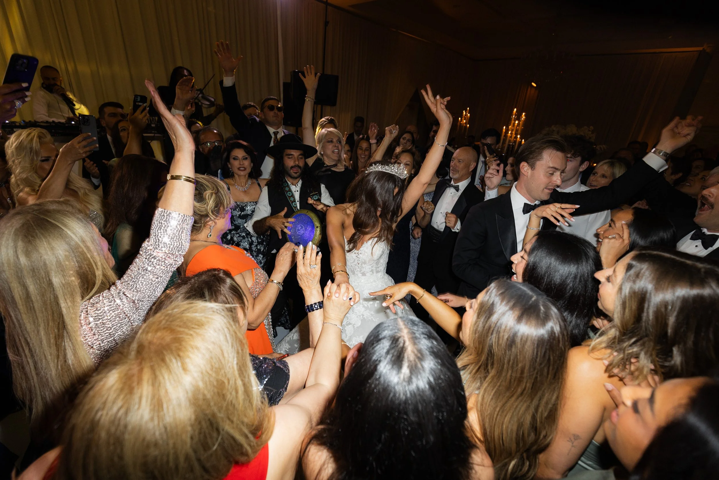 People dancing at a wedding reception, with a bride in a white gown surrounded by guests in formal attire, having a lively celebration.