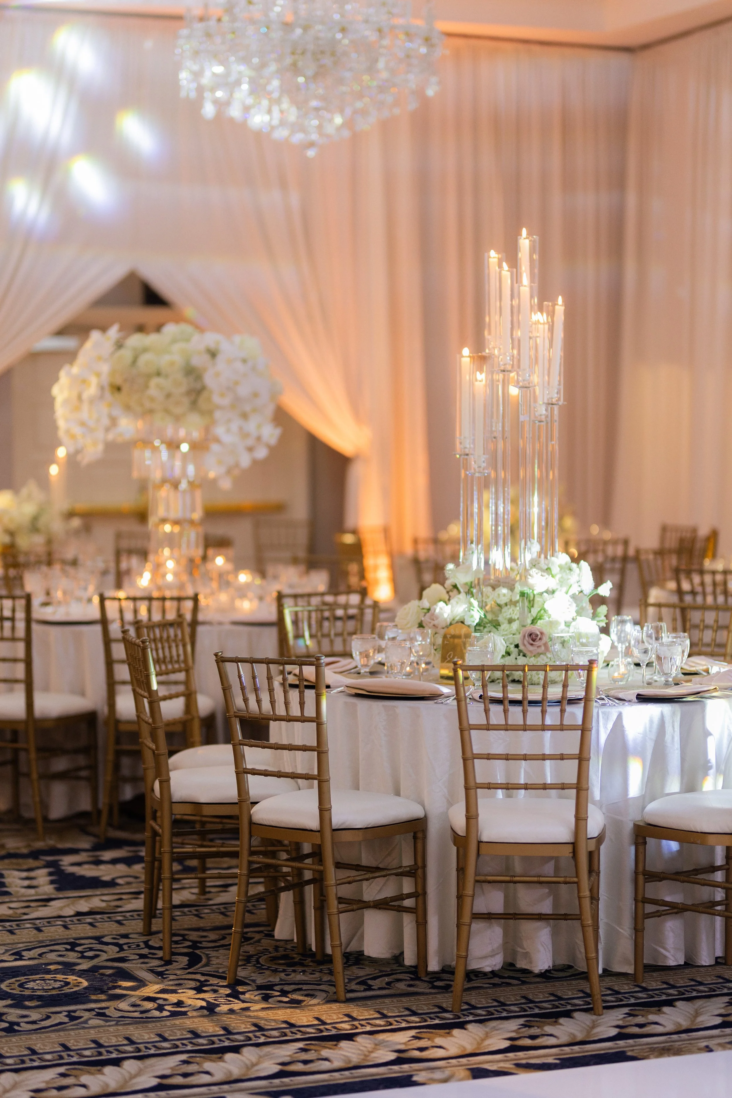 Elegant wedding reception setup with round tables, white tablecloths, tall floral centerpieces, gold chairs, crystal candelabras, and a chandelier.