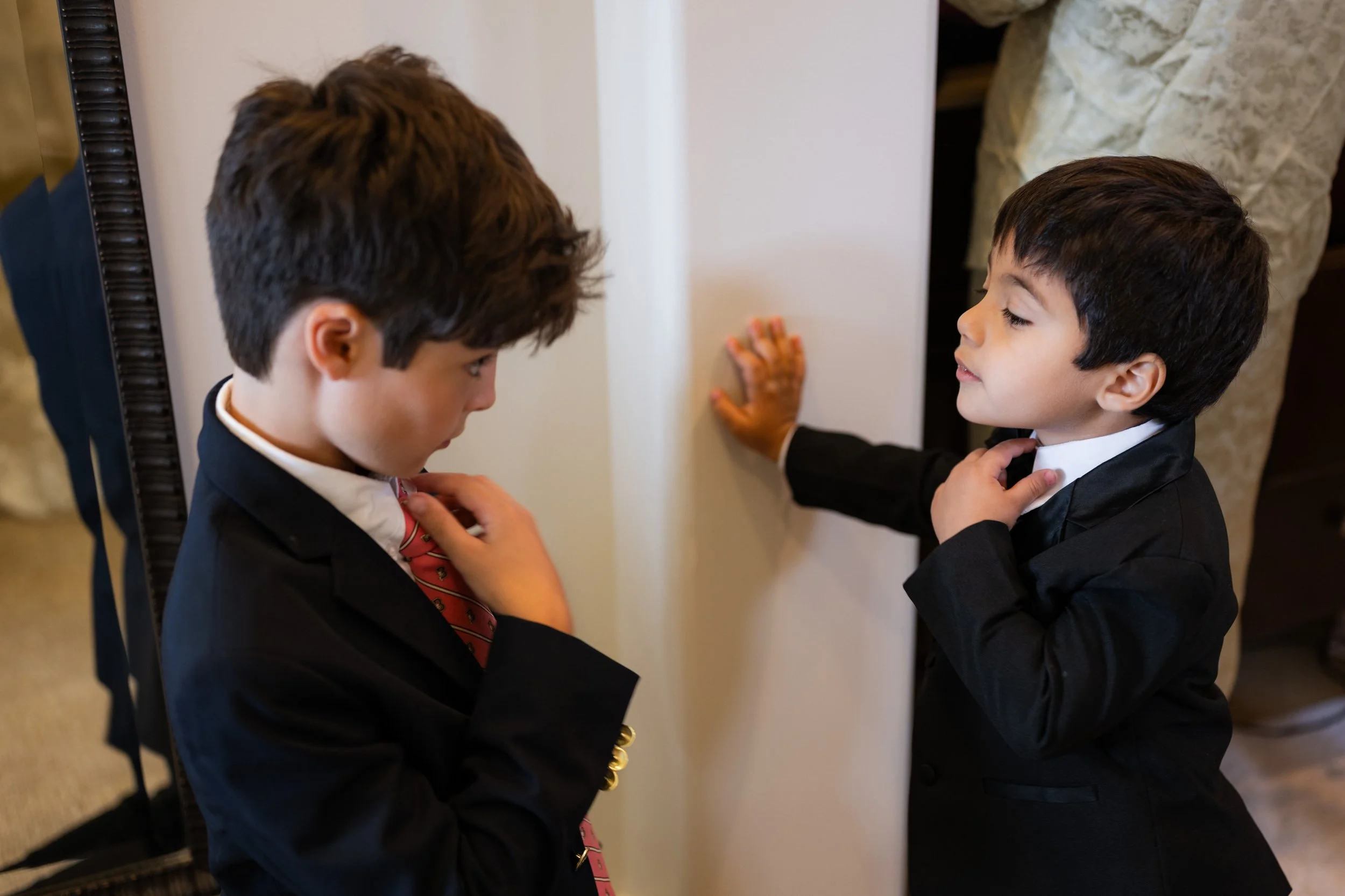 Two young boys in suits adjusting their ties, standing near a mirror, preparing for a formal event.