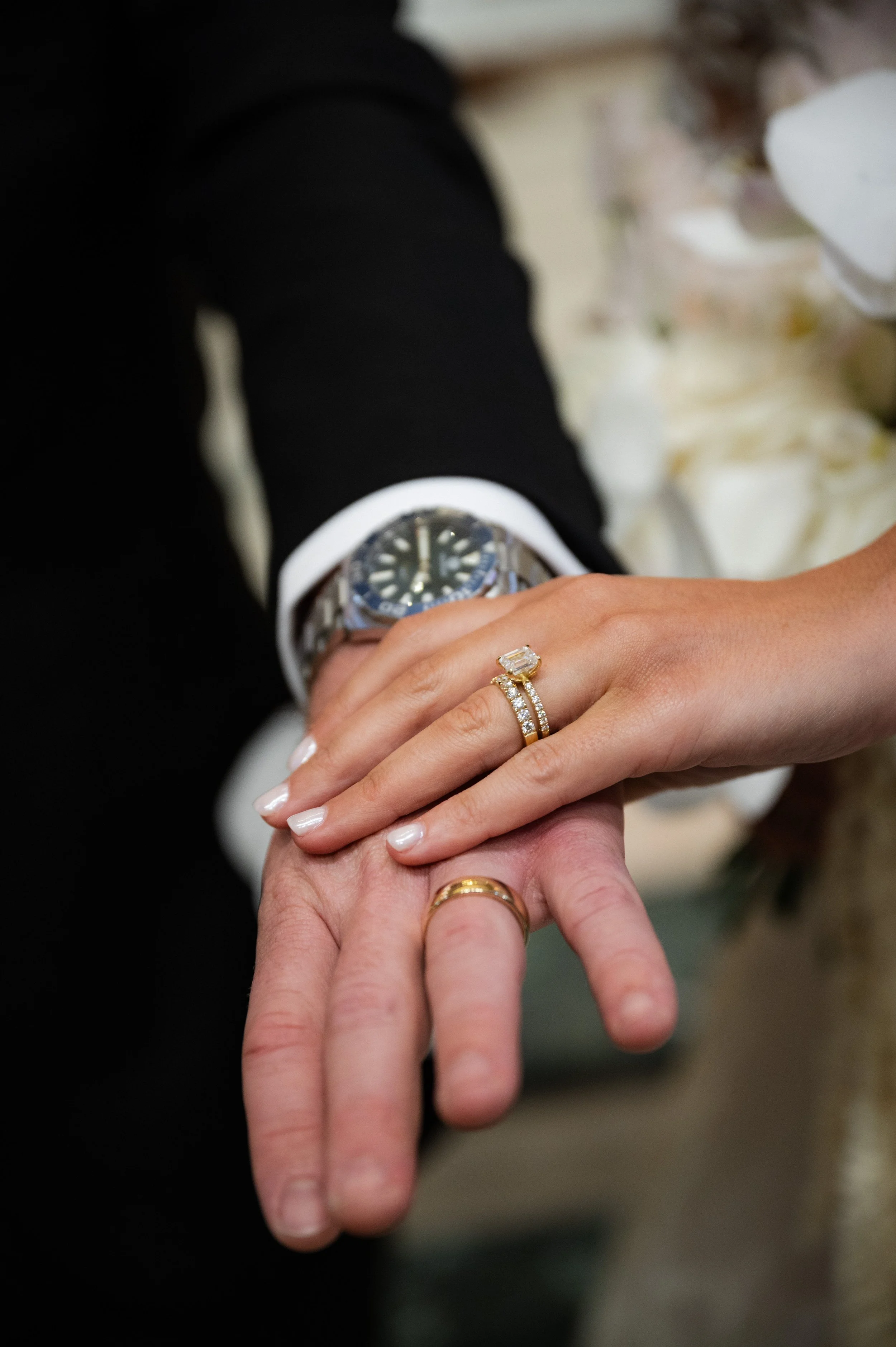 Close-up of a couple's hands showing wedding rings, with a man's hand in a suit and watch, and a woman's hand with an engagement and wedding ring.