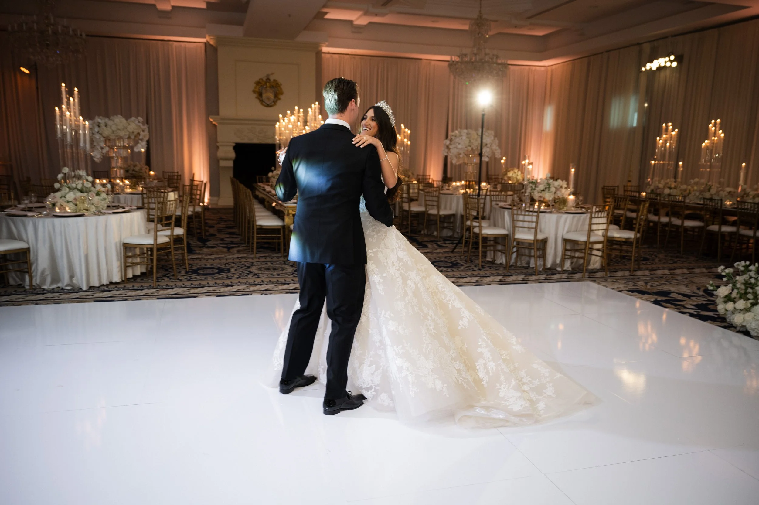 Bride and groom dancing in an elegant wedding reception hall with candlelit tables and floral decorations.