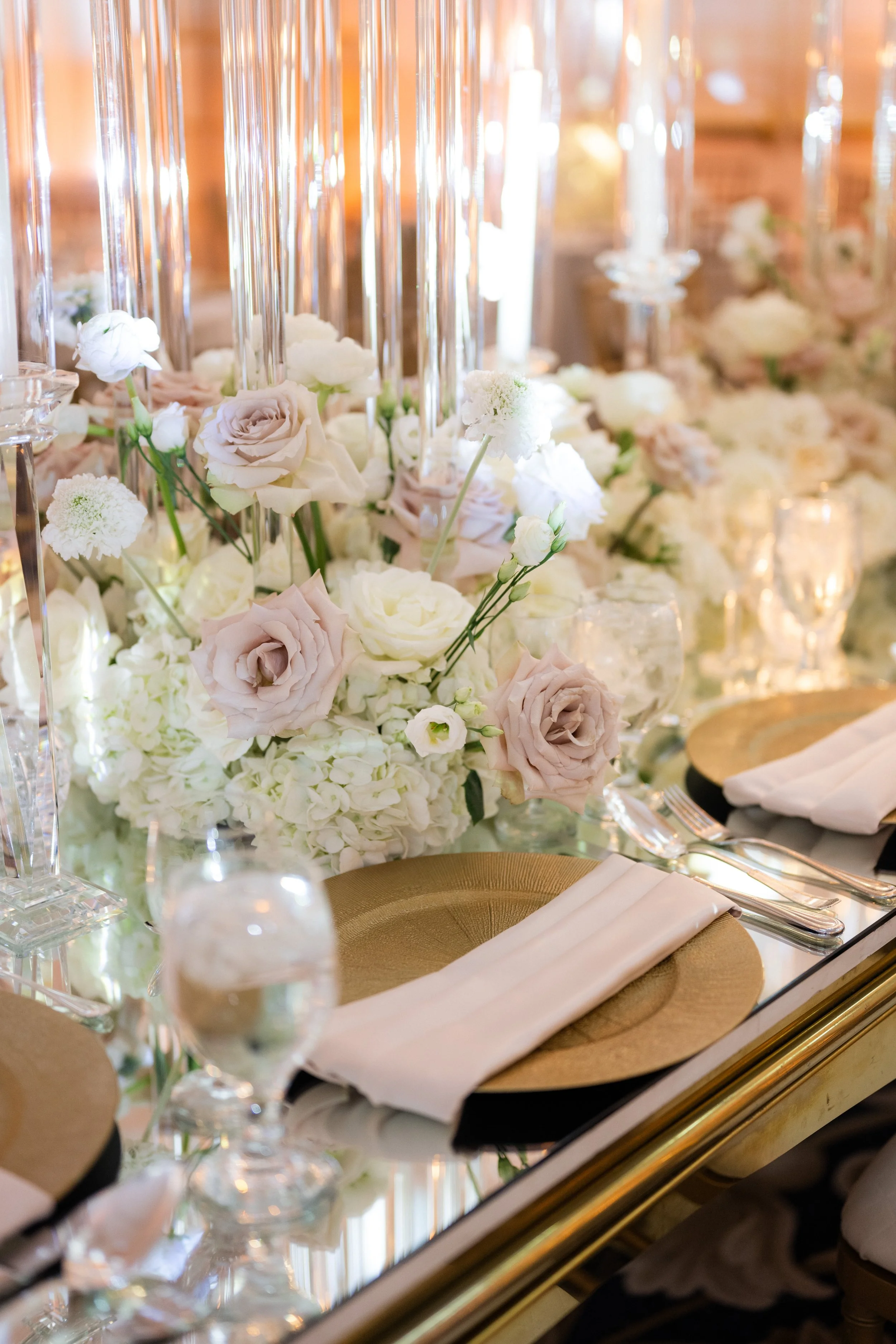Elegant table setting with gold plates, white napkins, crystal glasses, and a floral centerpiece featuring white and blush roses.