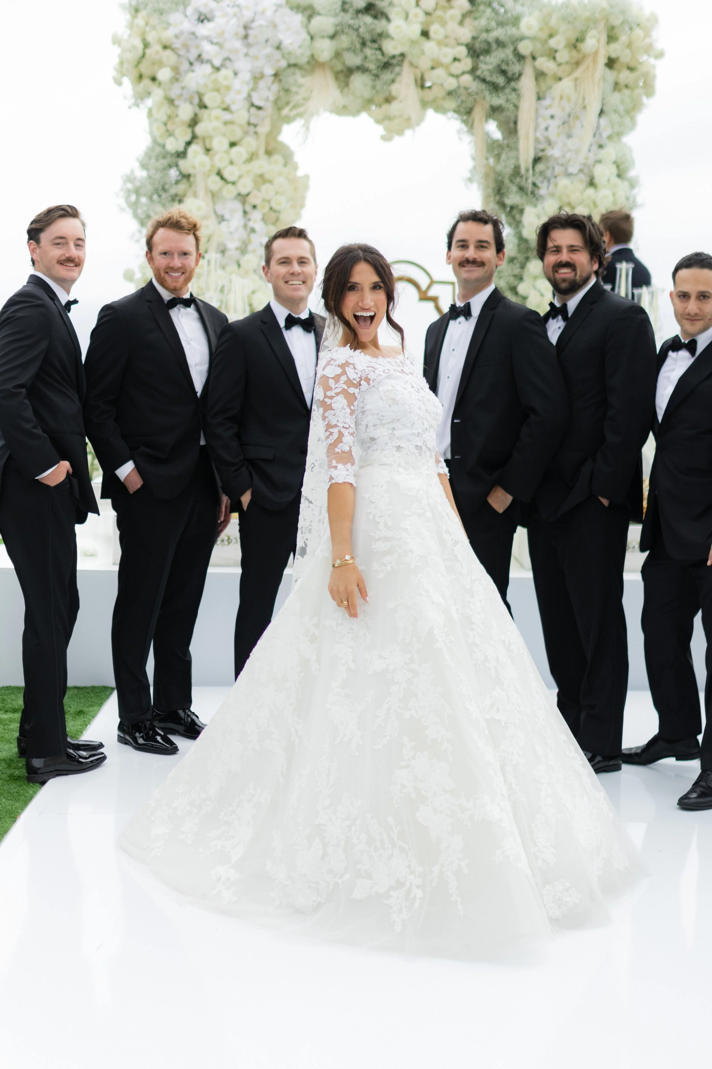 Bride in white lace dress poses with groomsmen in tuxedos, in front of floral arch at wedding.