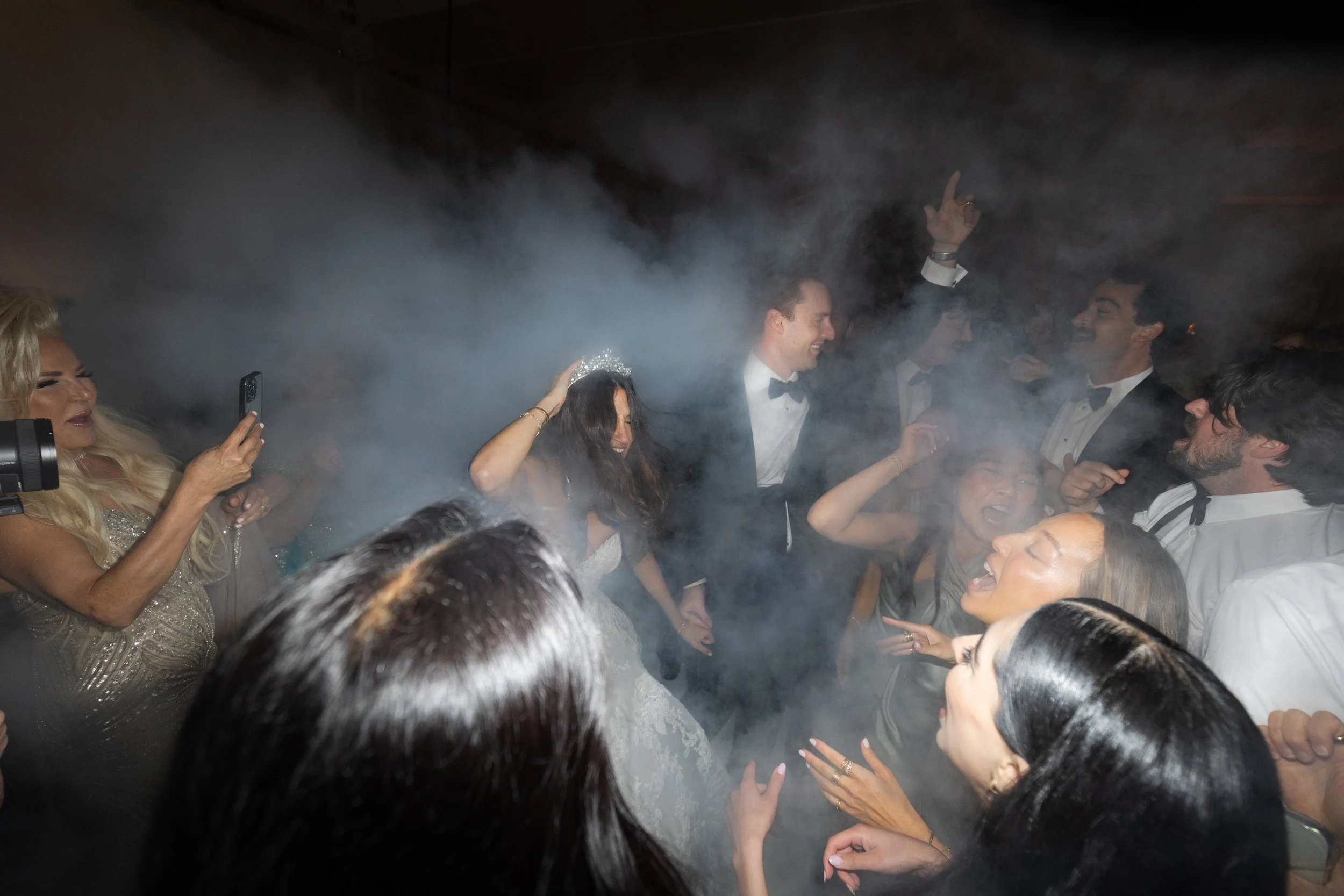People dancing in formal attire surrounded by fog or smoke, with one person taking a photo. The group appears to be celebrating or at a party.
