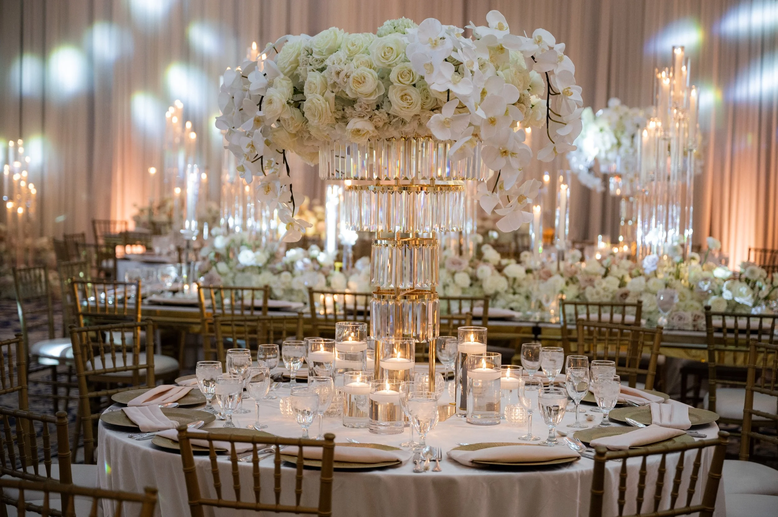 Elegant wedding reception table setup with white floral centerpiece, candles, gold chairs, and dim lighting.