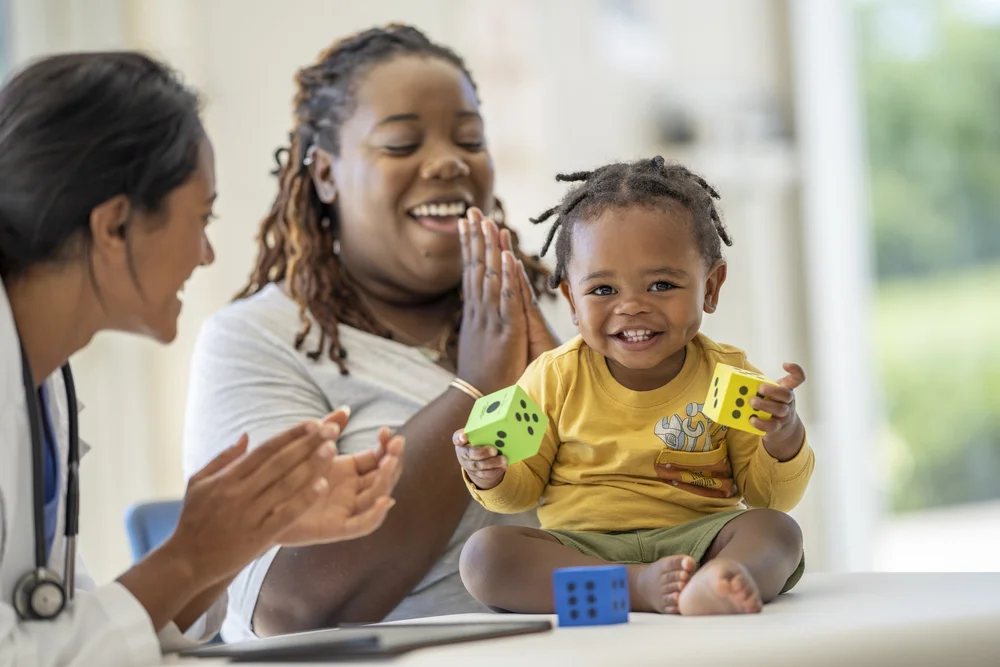 A caregiver playing with a smiling toddler while his mother looks on