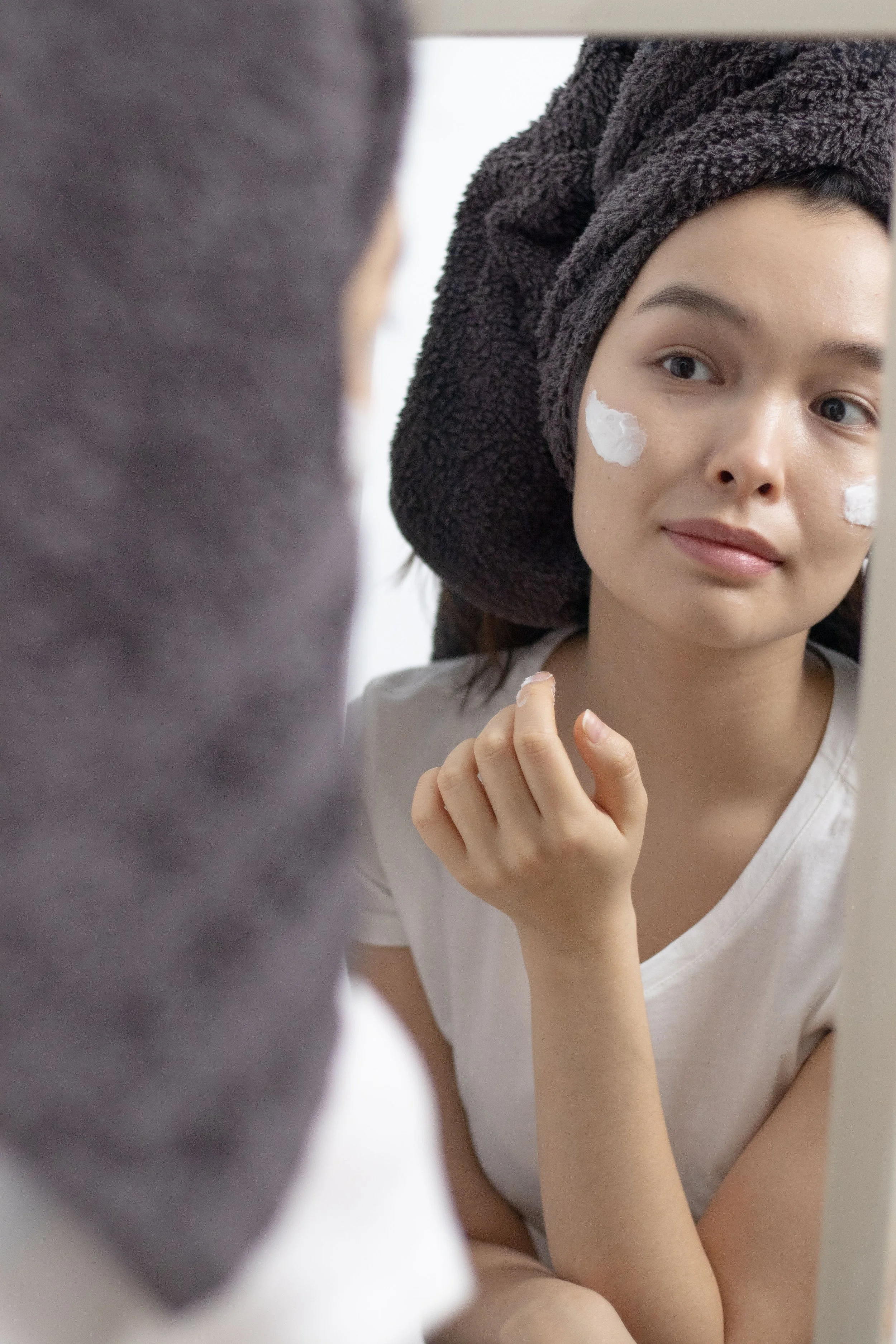 Young woman with a towel on her head applying cream to her cheek