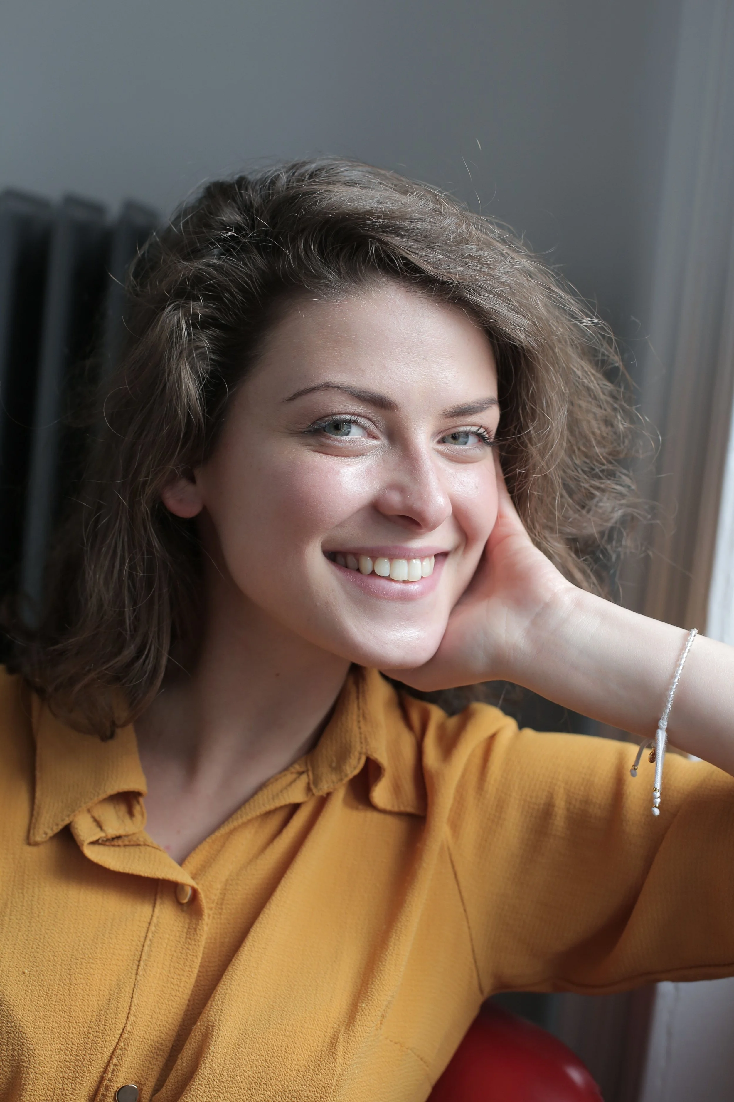 Smiling woman with curly brown hair in a mustard shirt, resting her hand against her cheek