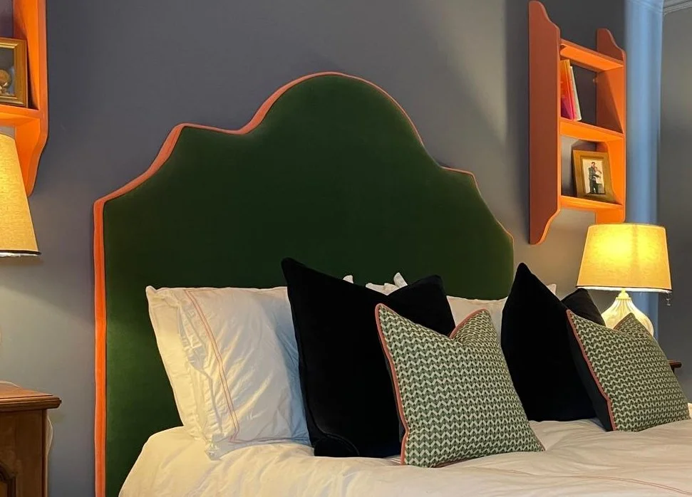A bedroom featuring a large green headboard with a scalloped orange border, two white pillows, and three decorative pillows with a chevron pattern, flanked by two lamps with yellow shades and orange wall-mounted shelves on a blue wall.