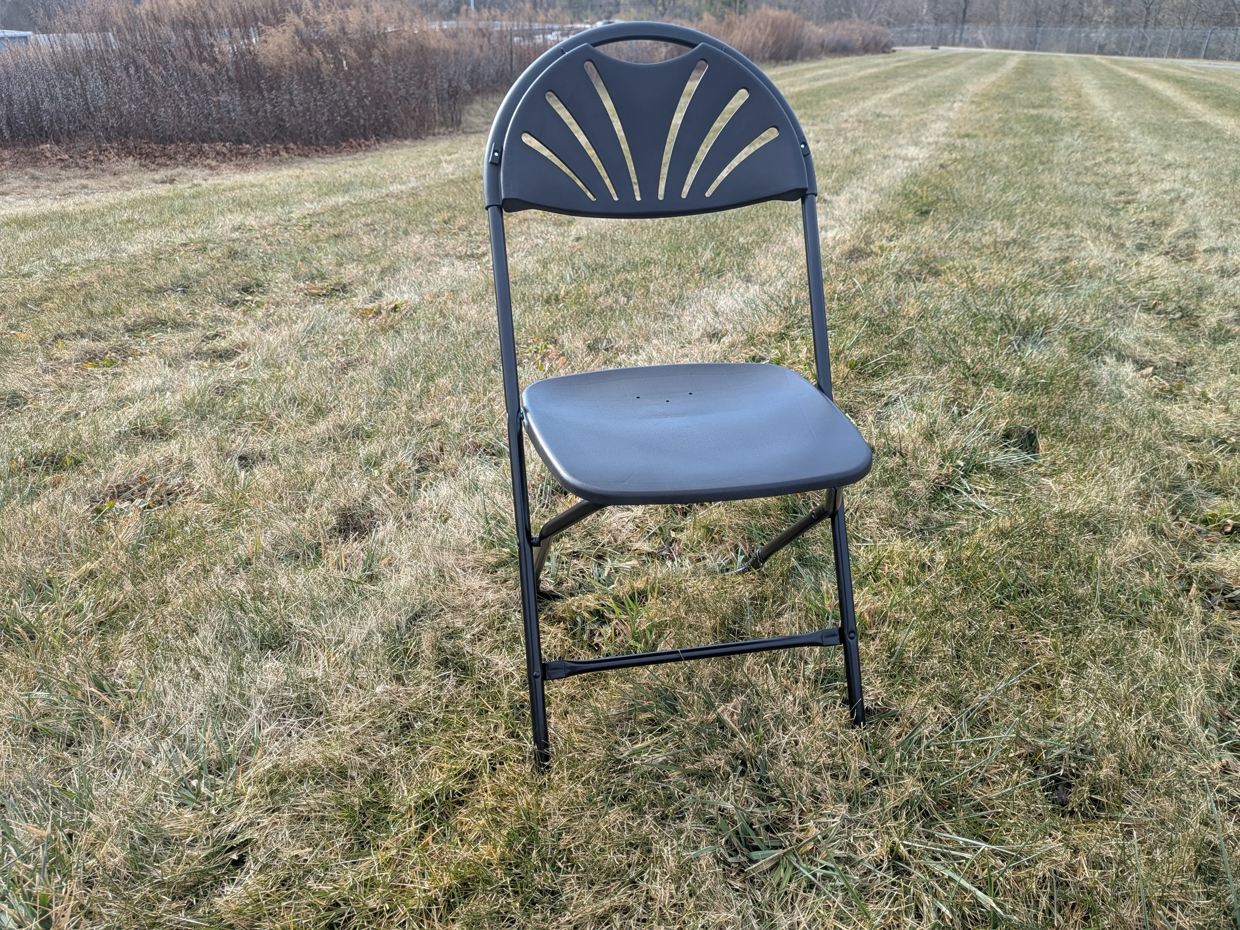 A black folding chair placed on grassy field with a hedge and trees in the background.