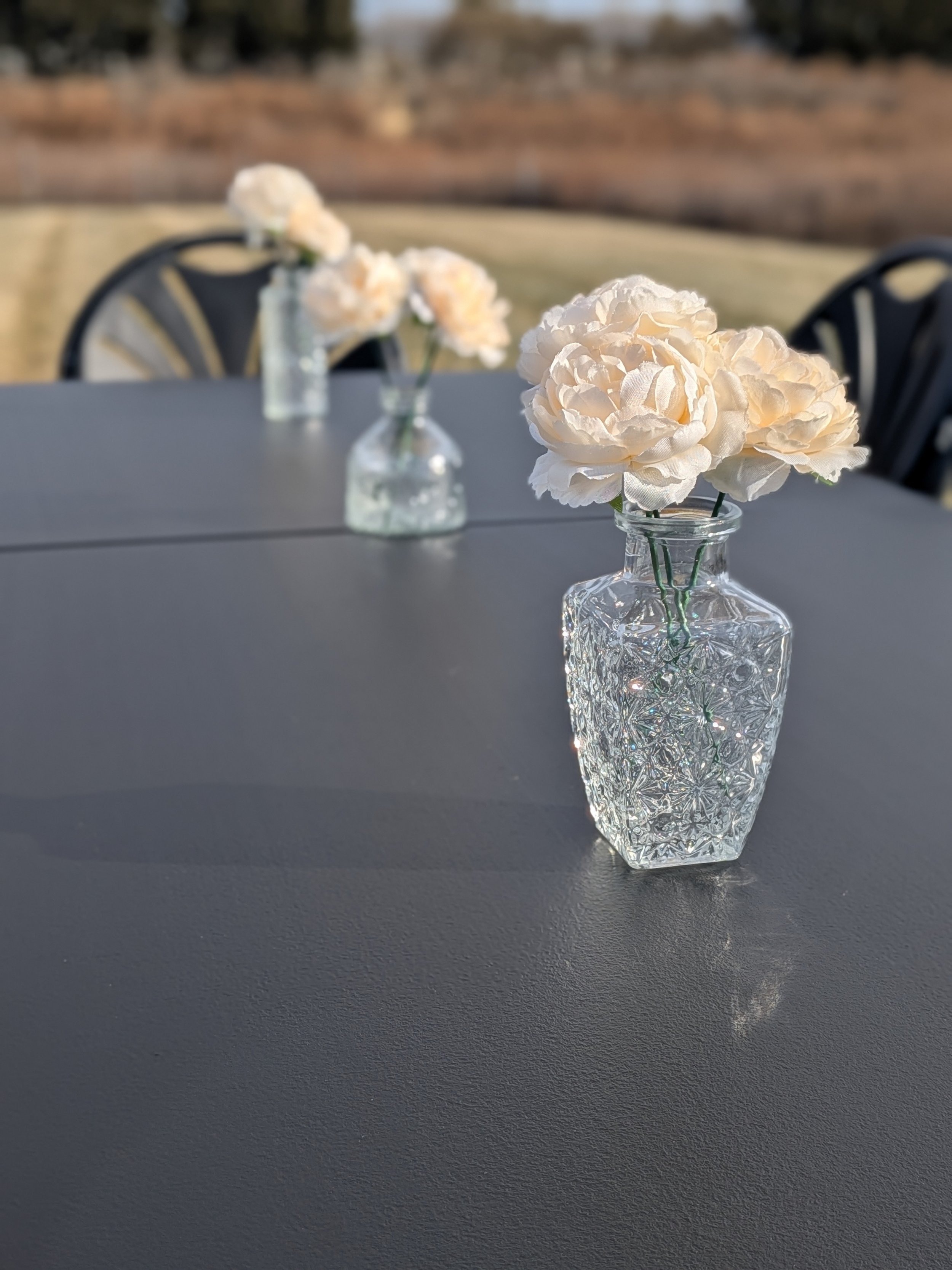 A close-up of a clear, patterned glass vase with white artificial flowers on a dark outdoor table, with more vases and flowers blurred in the background.