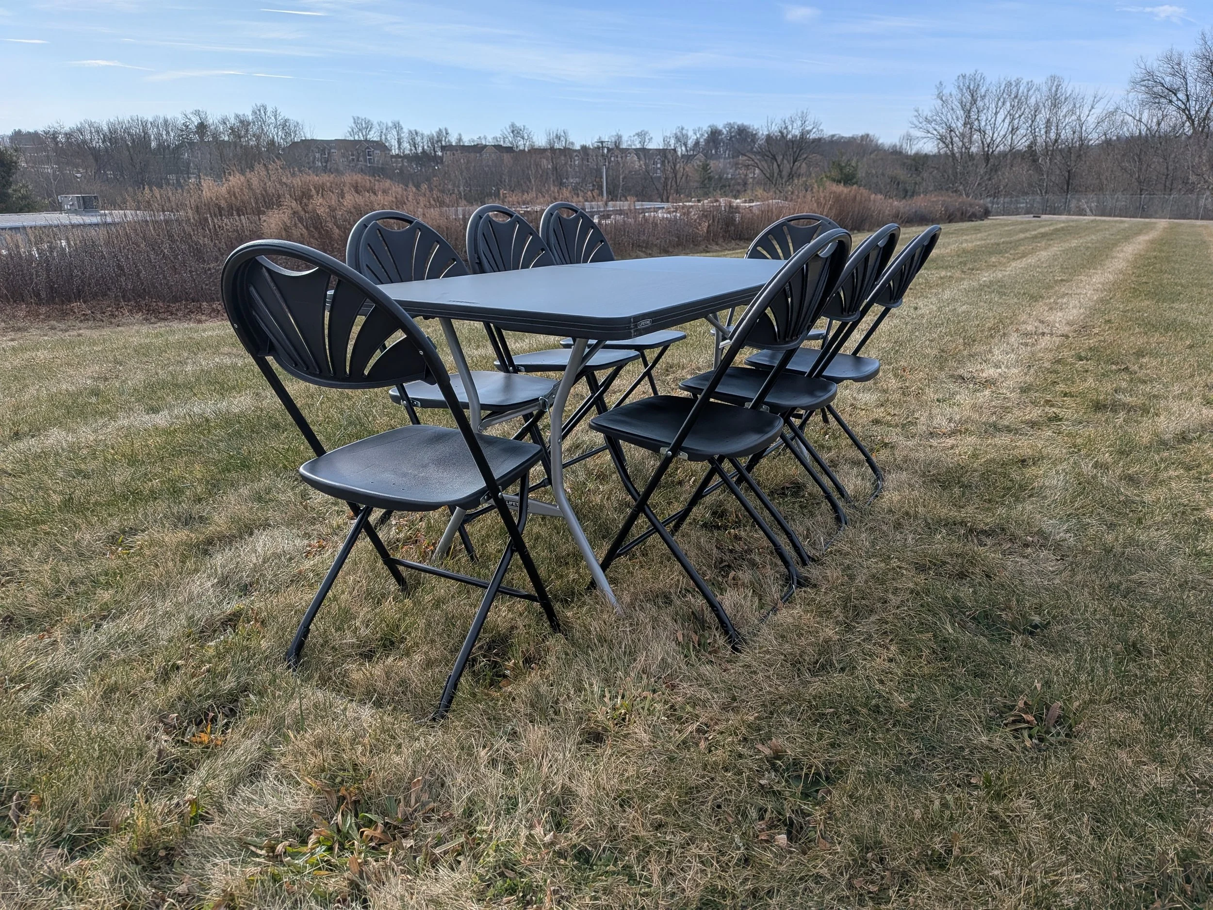 Outdoors, a black plastic table with eight matching chairs on a grassy field with bare trees and homes in the background under a partly cloudy sky.