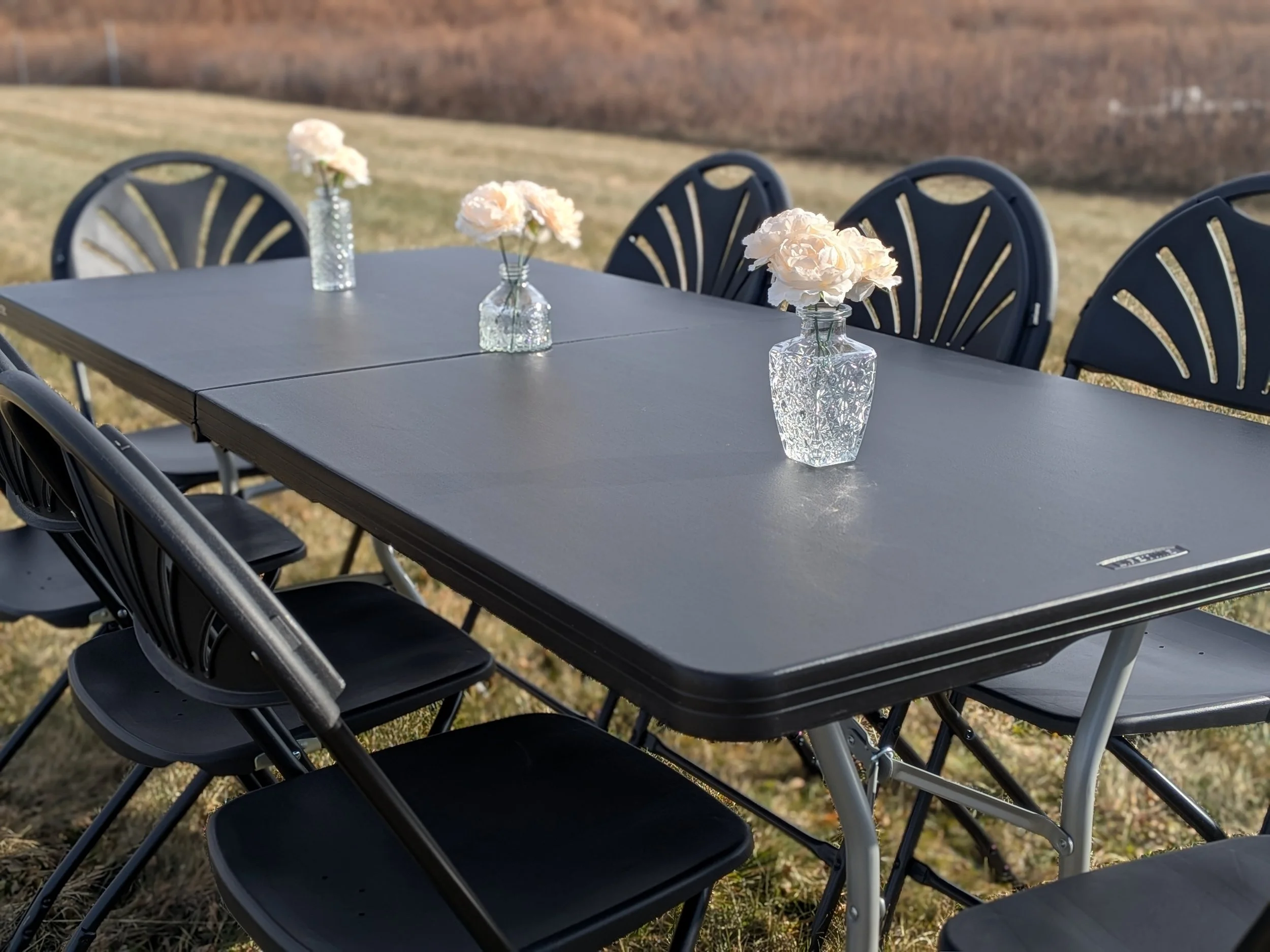 Outdoor dining table set with four vases of pink flowers, surrounded by black folding chairs, on grass with a blurry background of a fence and trees.
