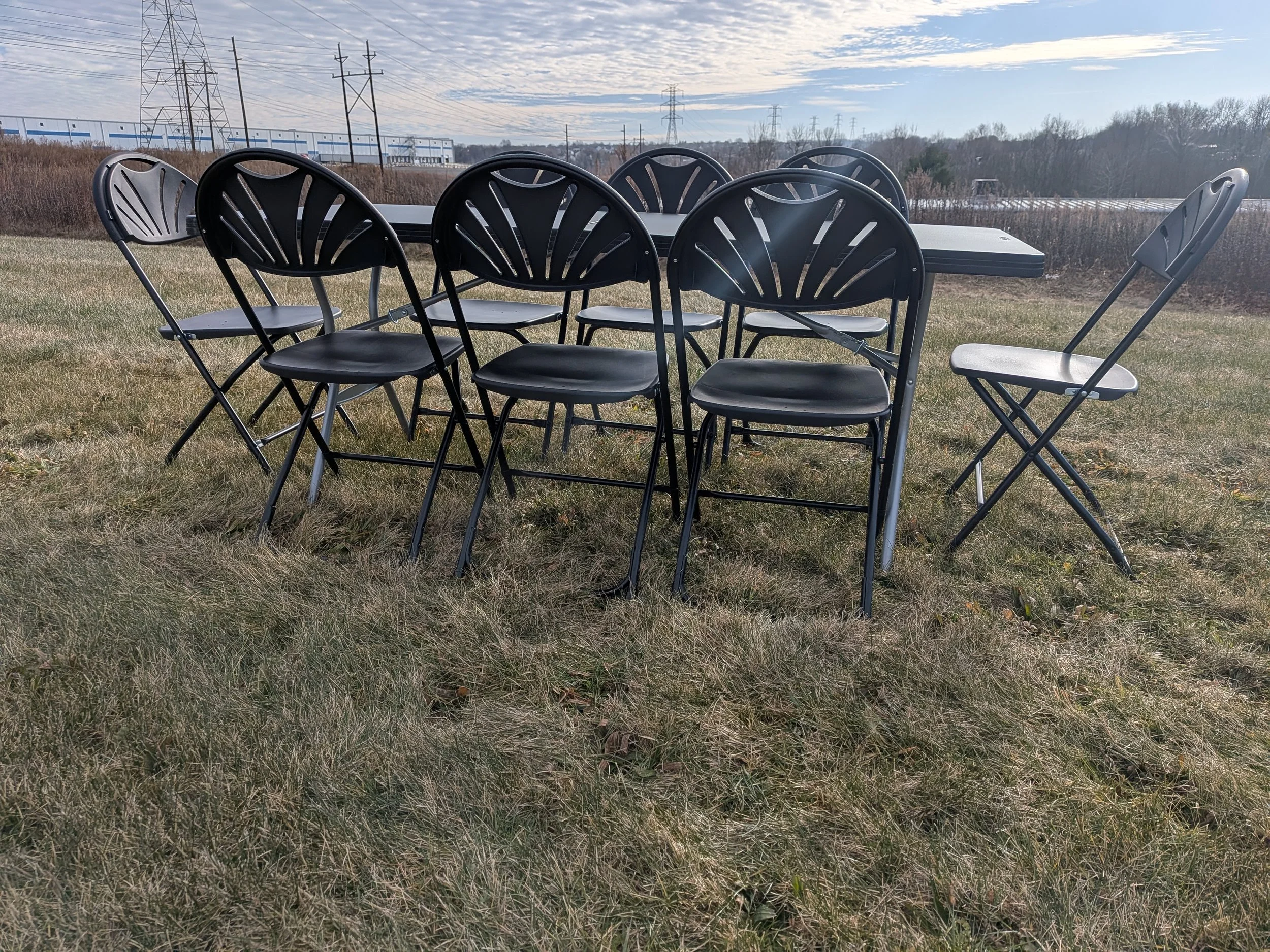 Eight black and gray folding chairs arranged around a black table outdoors, on a grassy field with power lines and industrial buildings in the background.