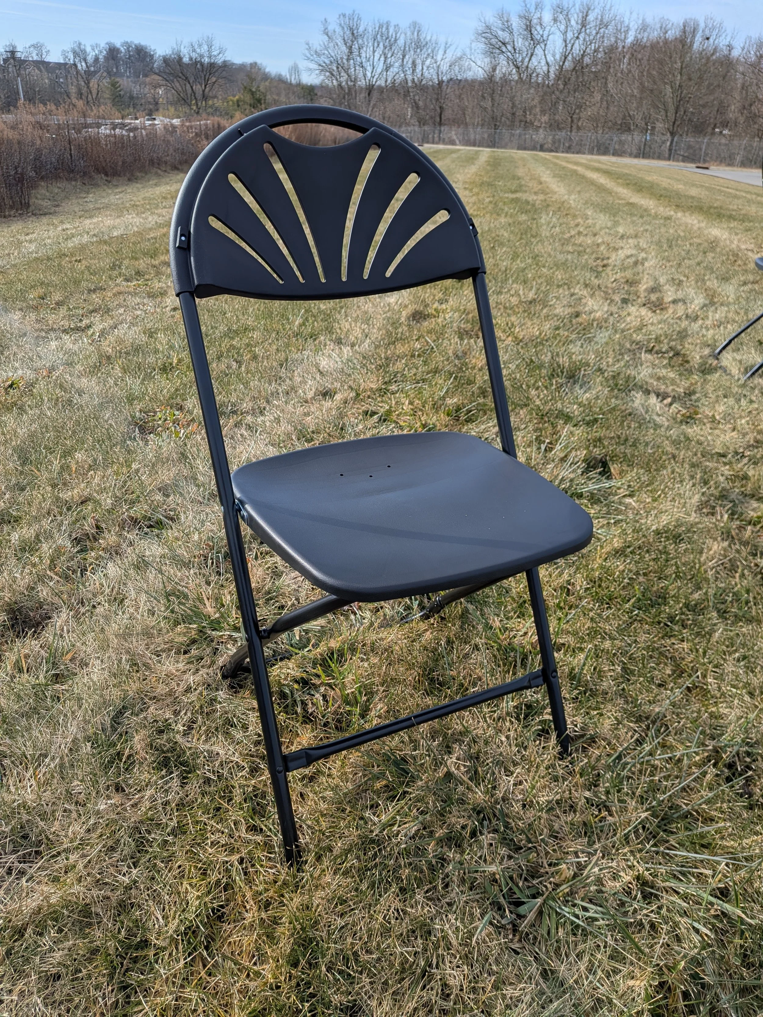Black folding chair outdoors on grassy field with trees and a road in the background.