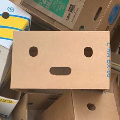 A cardboard box with a face design resembling a robot or character, with two circular cutouts for eyes and an oval cutout for a mouth, sitting among other boxes.