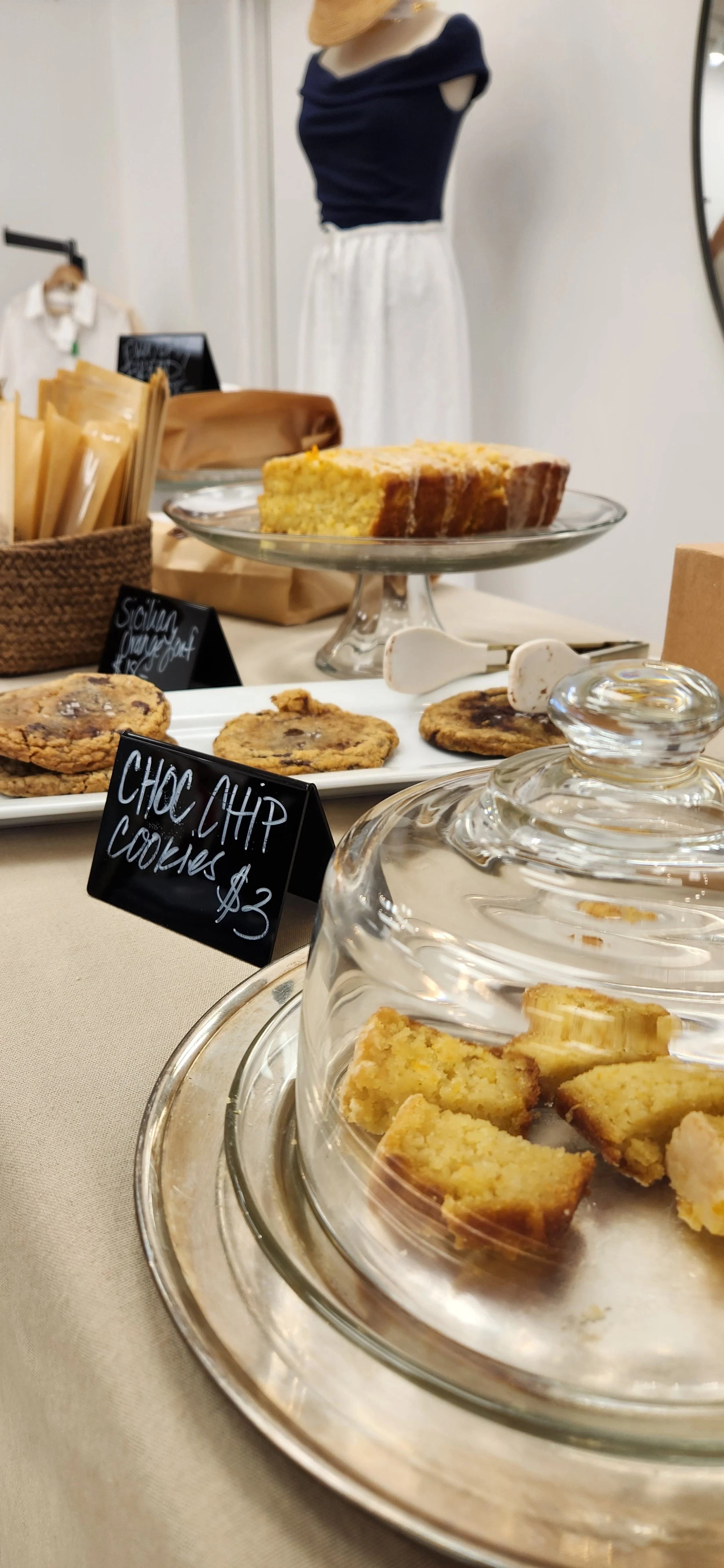 Display of chocolate chip cookies and lemon bar on plates and stands, with handwritten signs indicating prices, in a retail or bakery setting.