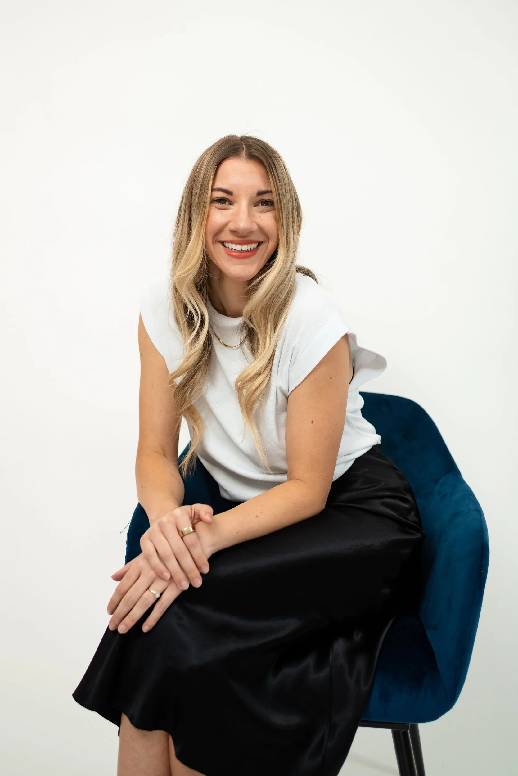 A photo of Brittany Bailey sitting in a blue velvet chair with a white blouse, black satin midi skirt with a gold chain.