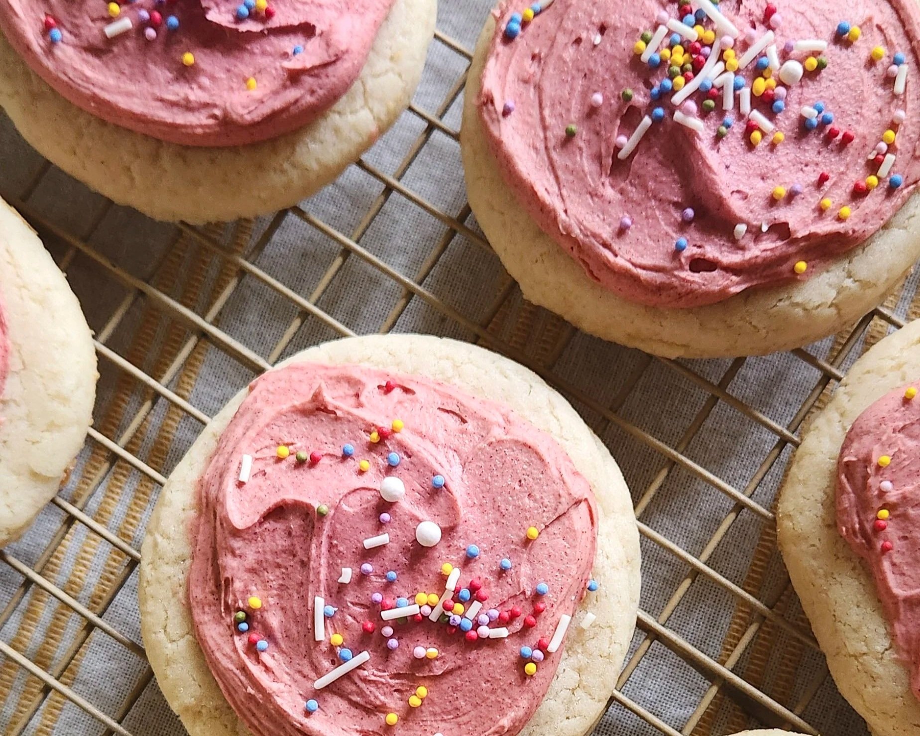 Close-up of an Ember and Bloom gluten-free and vegan Lofthouse sugar cookie featuring a thick layer of creamy pink frosting and colorful rainbow sprinkles on a soft, cake-like base.