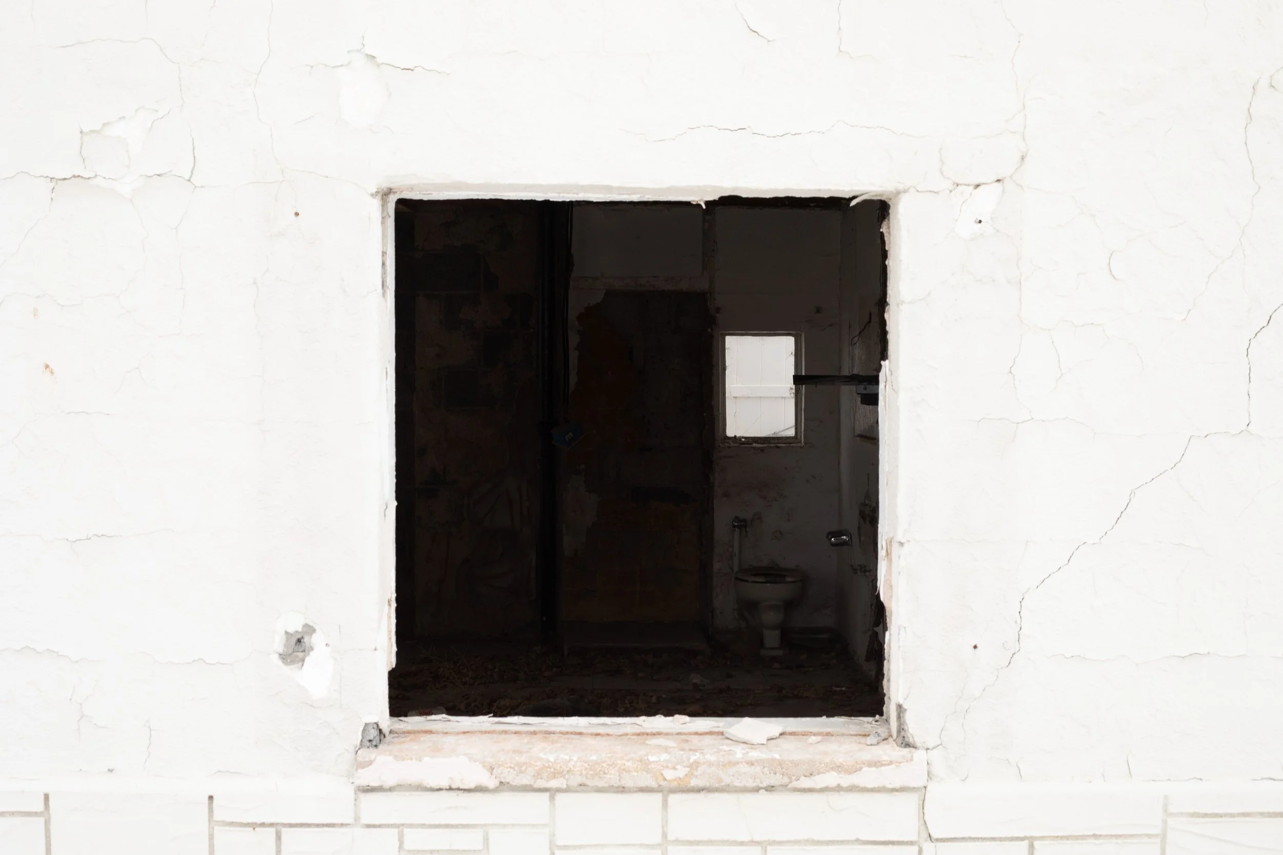 Square window opening in peeling white stucco, dark interior beyond, abandoned motel, Sterling, CO